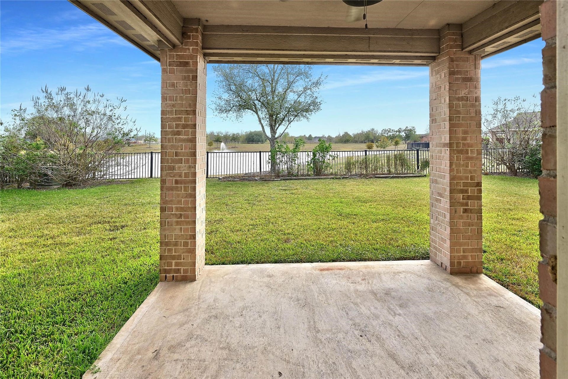 5936 Pearland Place, Pearland, TX 77581 This photo shows a covered patio with brick columns, overlooking a spacious backyard. The area is grassy and fenced, offering a view of a serene pond with a fountain and surrounding greenery, perfect for relaxation and outdoor enjoyment.