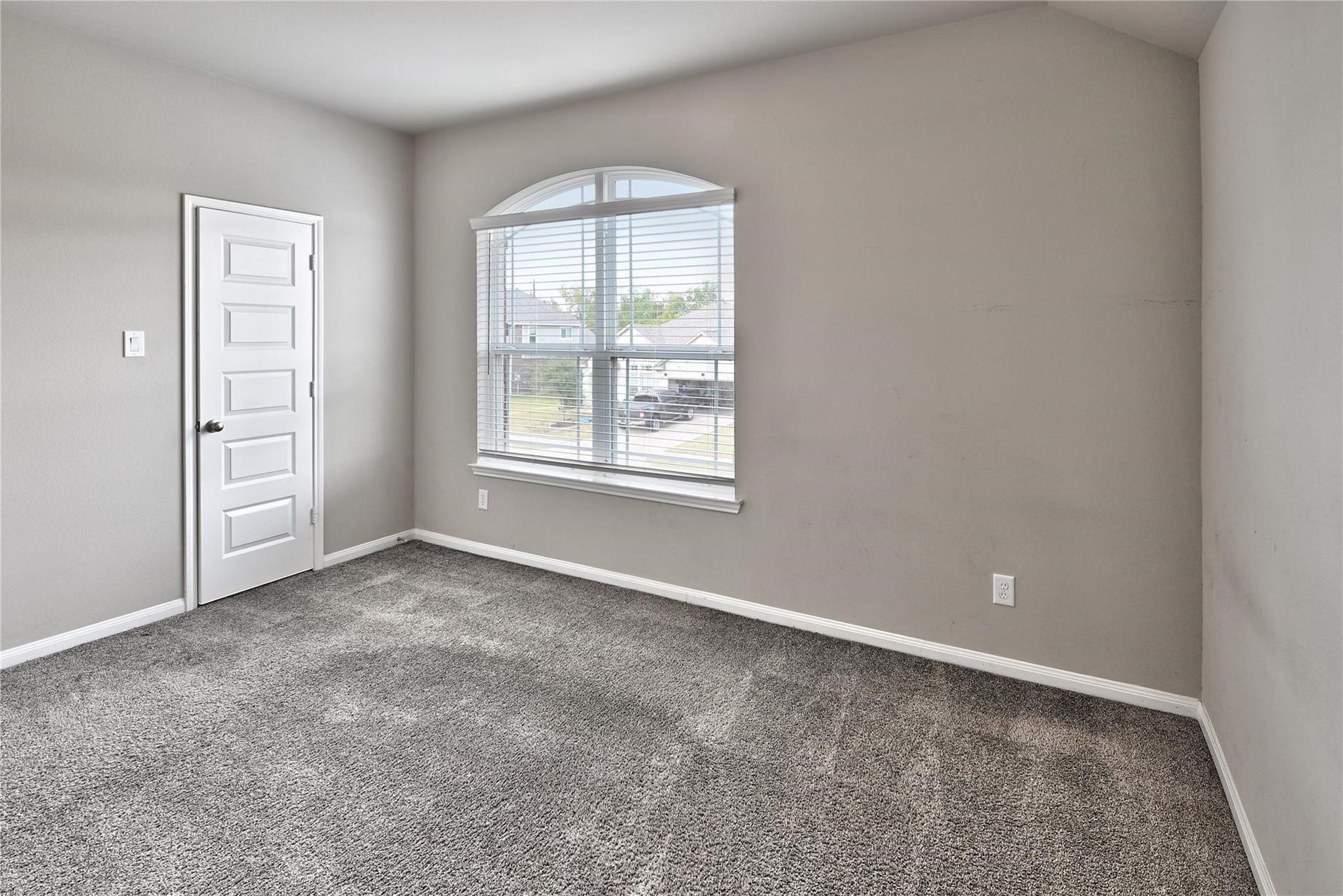 5936 Pearland Place, Pearland, TX 77581 The photo shows a bright, empty room with neutral walls and carpet flooring. It features a large window with blinds, allowing natural light, and a white paneled door. Perfect for a bedroom or office space.