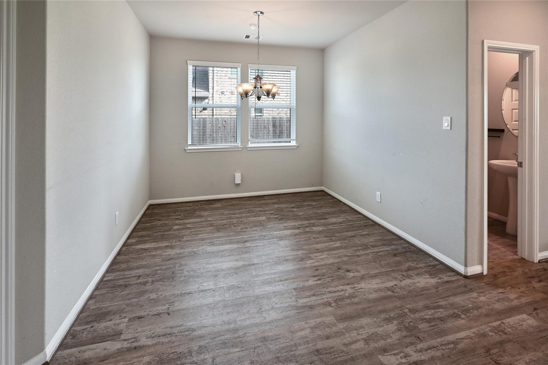 5936 Pearland Place, Pearland, TX 77581 This photo shows a bright, empty room with light gray walls and wood-style flooring, featuring a central light fixture and two windows. There is an adjoining small bathroom with a pedestal sink.