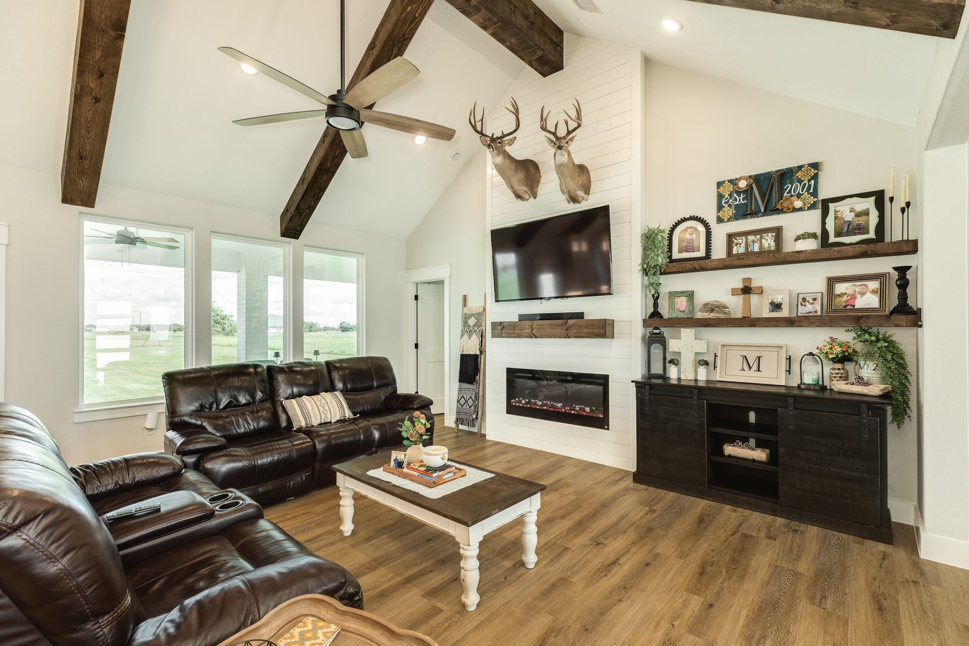 608 Angus Trail, Angleton, TX 77515 A stylish living room featuring a vaulted ceiling with exposed wood beams, and a cozy electric fireplace. Natural light floods through large windows, creating a warm and inviting atmosphere.