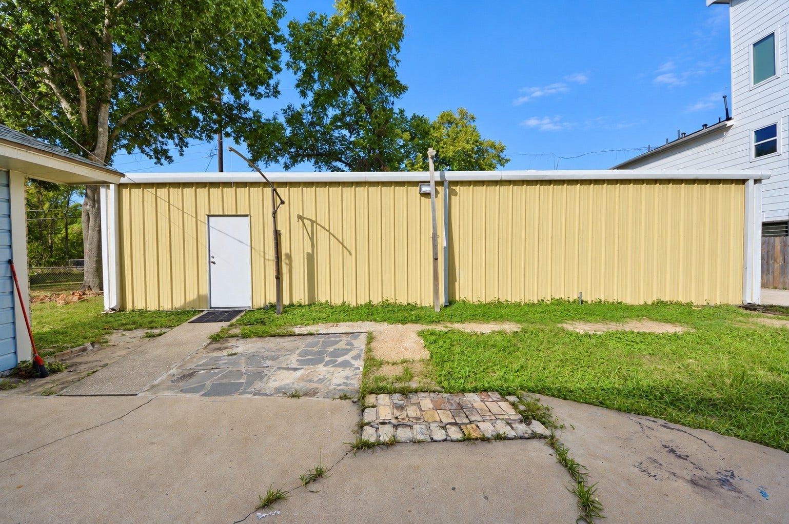 1837 Wycliffe Drive, Houston, TX 77043 A spacious backyard features a sizable yellow metal storage building with a white door, surrounded by a mix of concrete and grassy areas, ideal for additional storage or a workshop.