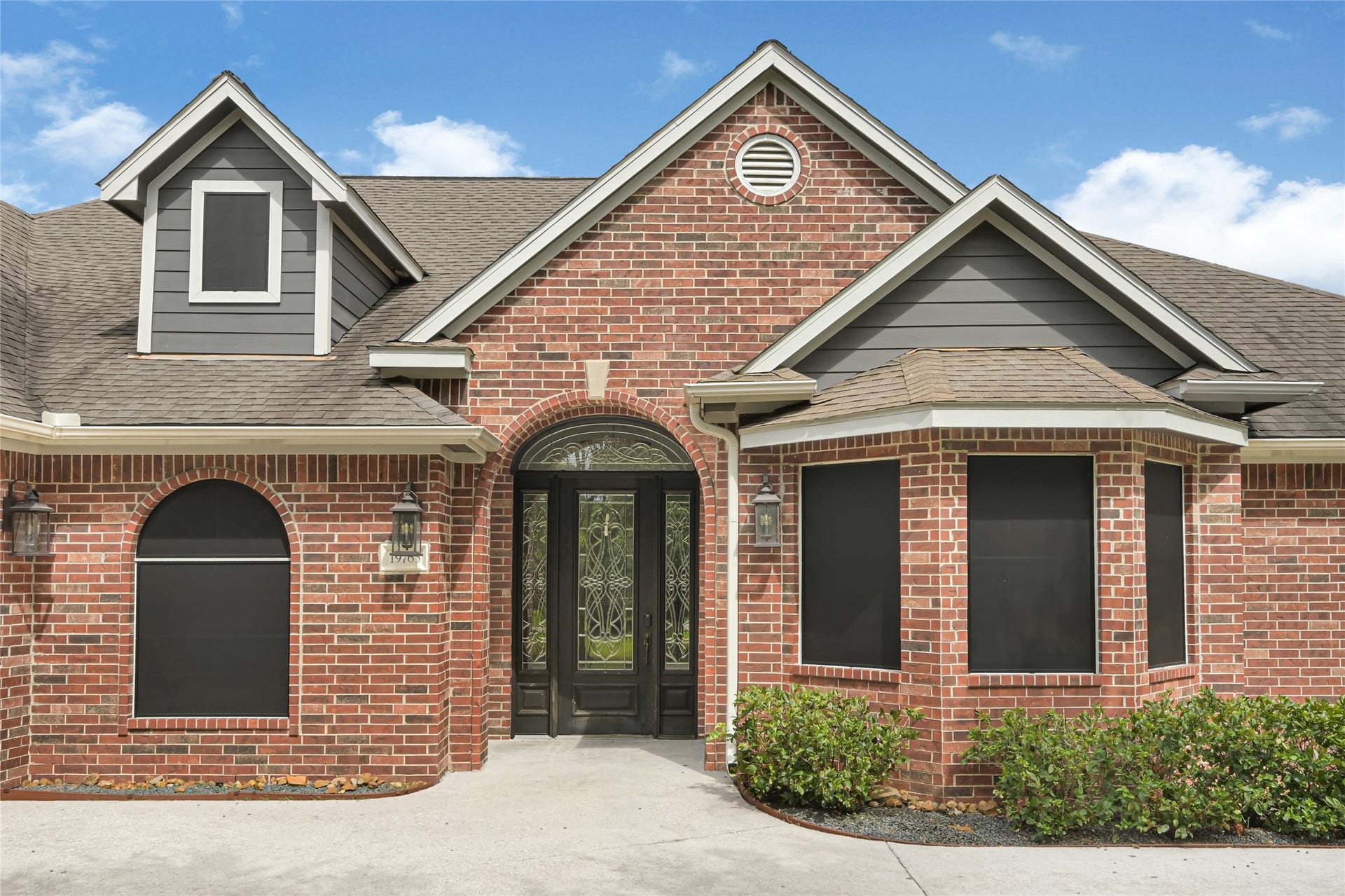 19763 Irenell Dr, Porter, TX 77365 Charming brick facade with elegant arched entry and decorative glass door, complemented by neatly manicured shrubs and stylish roof gables.