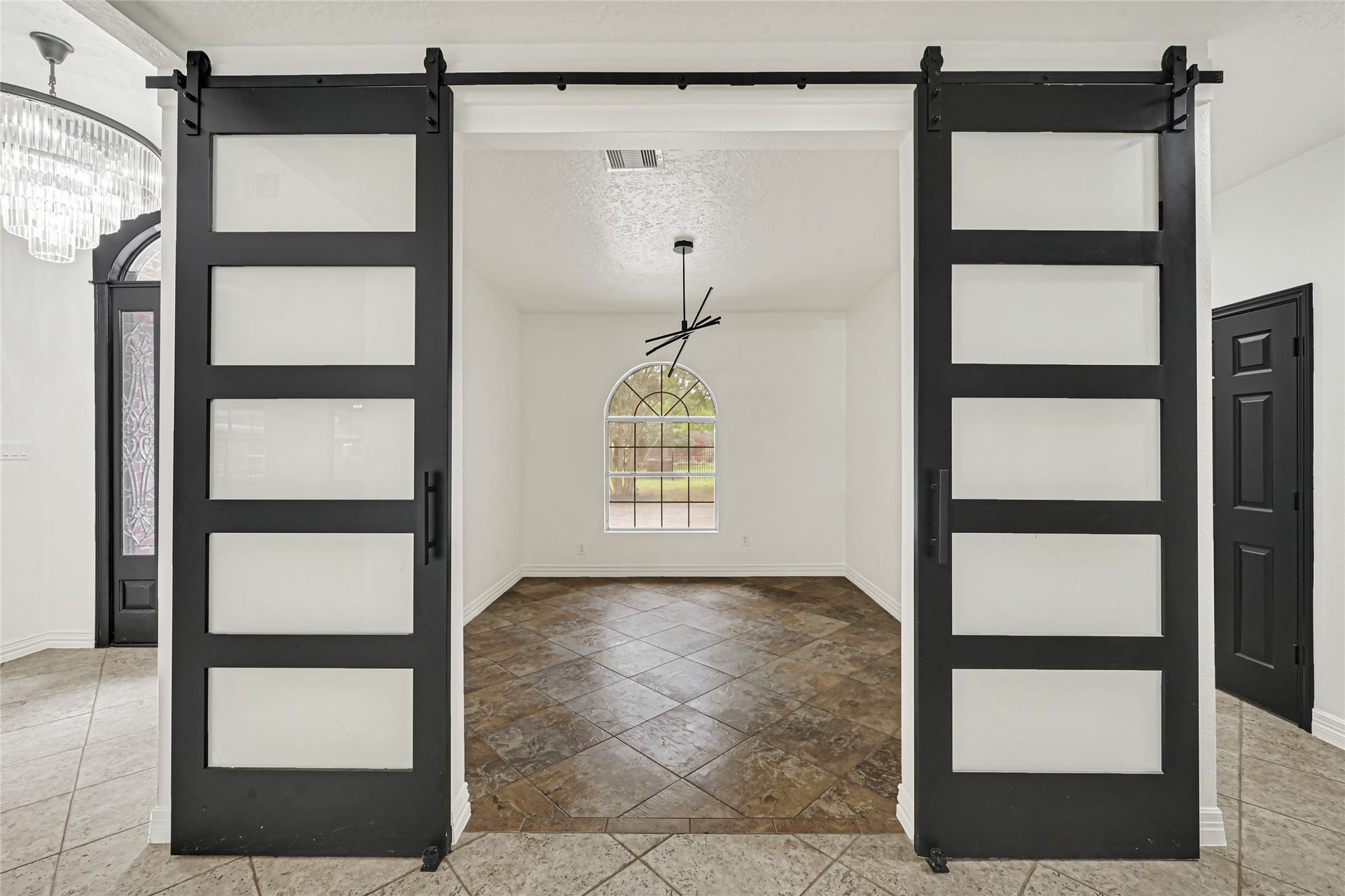 19763 Irenell Dr, Porter, TX 77365 Sleek black sliding barn doors lead into a bright room with a modern chandelier and elegant arched window, featuring stylish tile flooring.