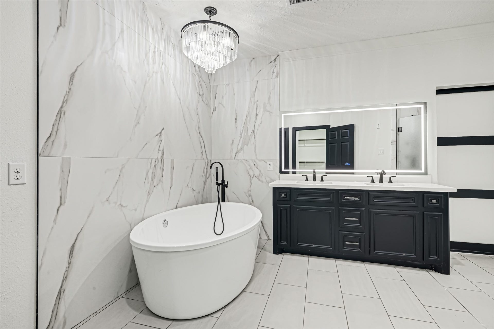 19763 Irenell Dr, Porter, TX 77365 Luxurious bathroom featuring a sleek freestanding bathtub, elegant marble walls, a modern double vanity with a backlit mirror, and a sophisticated chandelier.