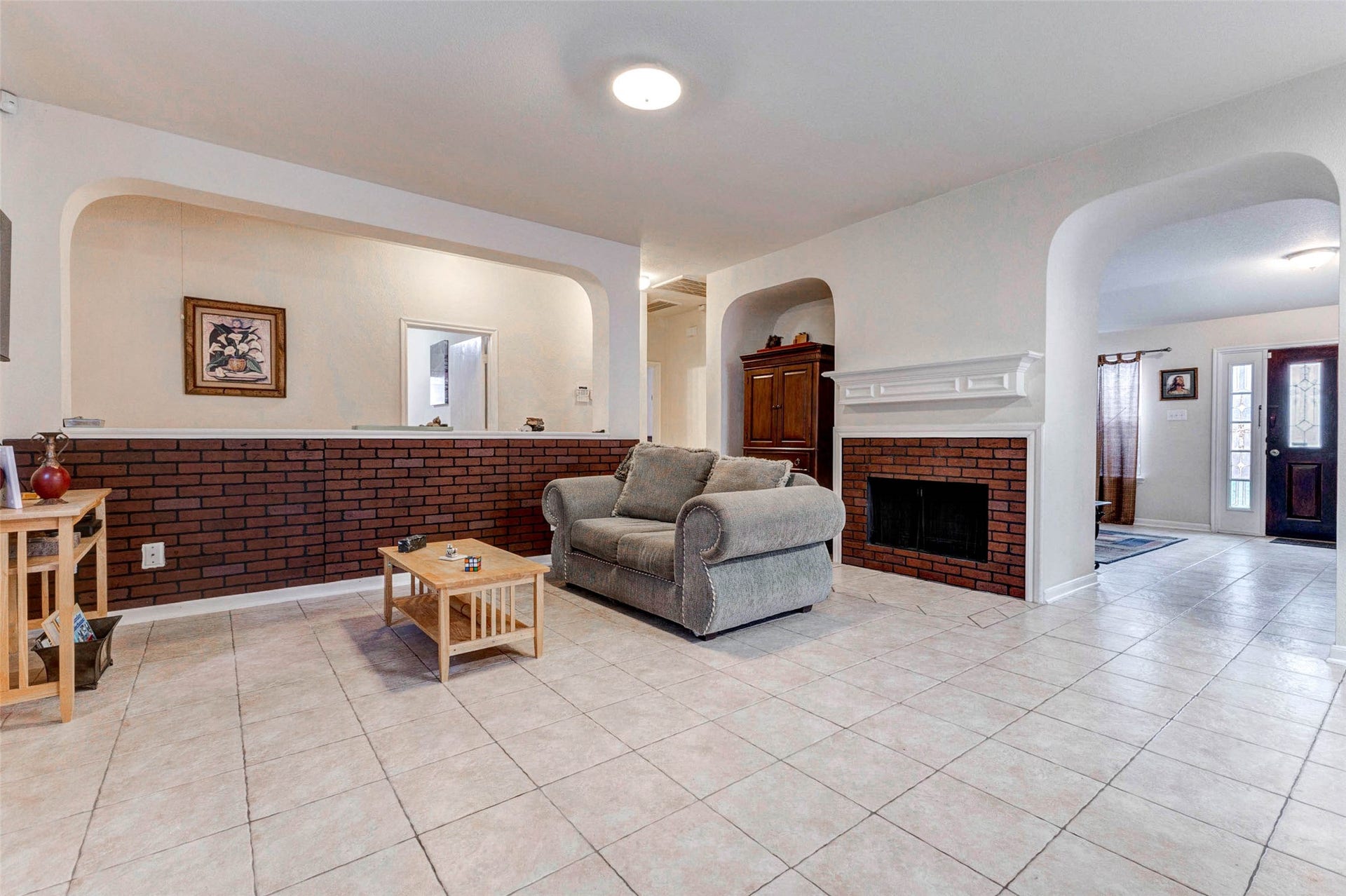 21211 Arcadia Park Lane, Humble, TX 77338 Spacious living area featuring an open layout with durable tile flooring and a cozy brick-accent fireplace that adds warmth and character. Arched openings and a half-wall design create visual separation while maintaining an airy, connected feel. Neutral tones throughout provide a versatile backdrop, while natural light enhances the inviting atmosphere, making this space perfect for relaxing or entertaining guests.