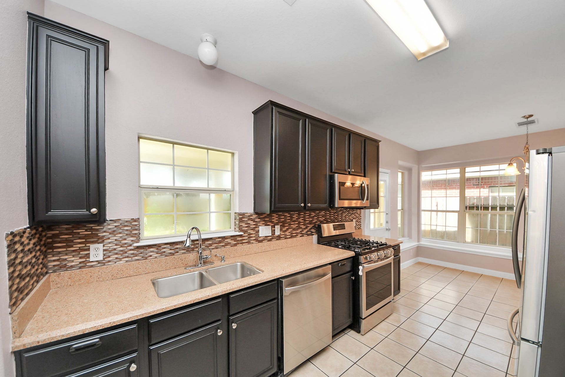10026 Gil Jr Lane, Houston, TX 77075 Modern kitchen with sleek dark cabinets, stainless steel appliances, and a stylish backsplash. Ample natural light streams through large windows, highlighting the spacious, tiled floor.