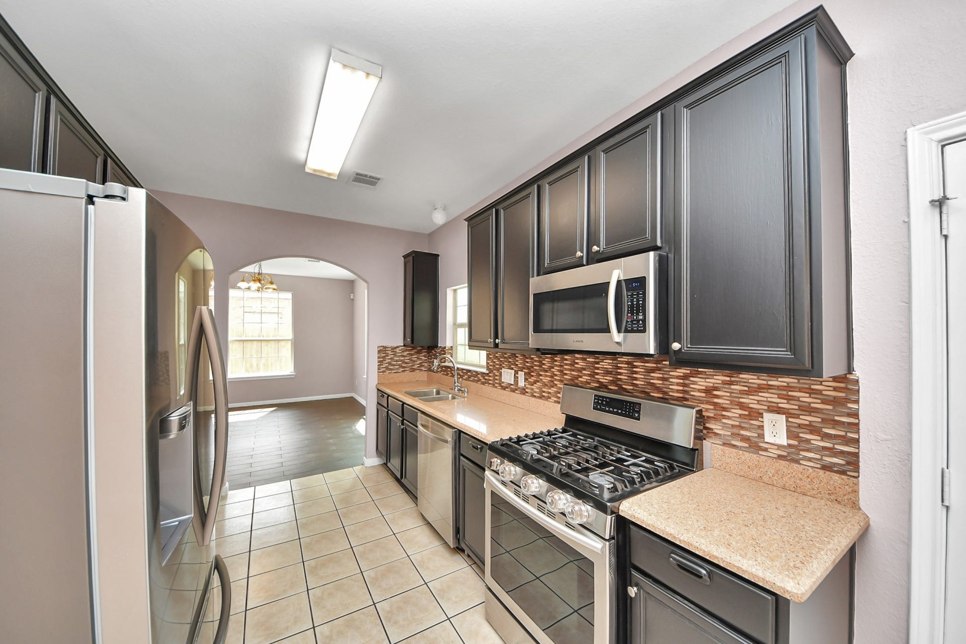 10026 Gil Jr Lane, Houston, TX 77075 Sleek kitchen featuring dark cabinetry, modern stainless steel appliances, and a stylish backsplash, leading into a bright dining area with large windows.