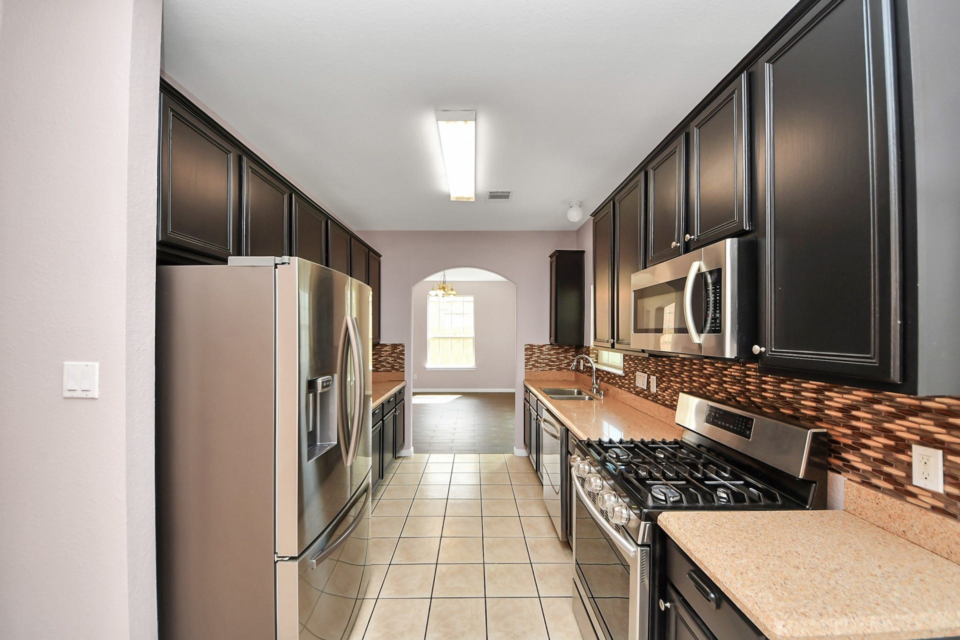10026 Gil Jr Lane, Houston, TX 77075 Charming galley kitchen featuring sleek dark cabinetry, modern stainless steel appliances, and a stylish tile backsplash, leading to a cozy dining area filled with natural light.
