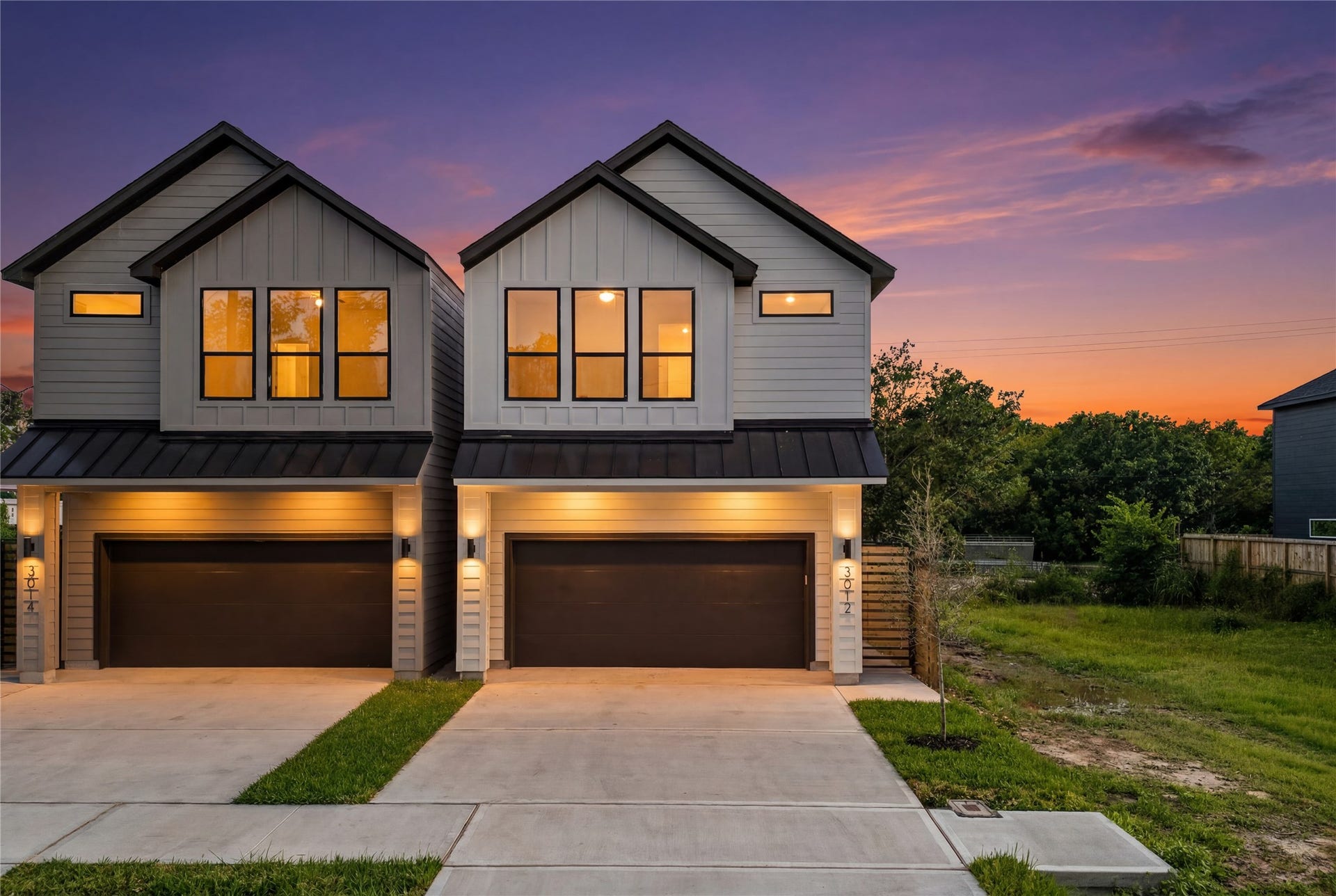 This close-up twilight view focuses on the sleek entryways and dark garage doors. Warm accent lighting glows from the large windows, highlighting the clean lines and modern design elements of the home's exterior.