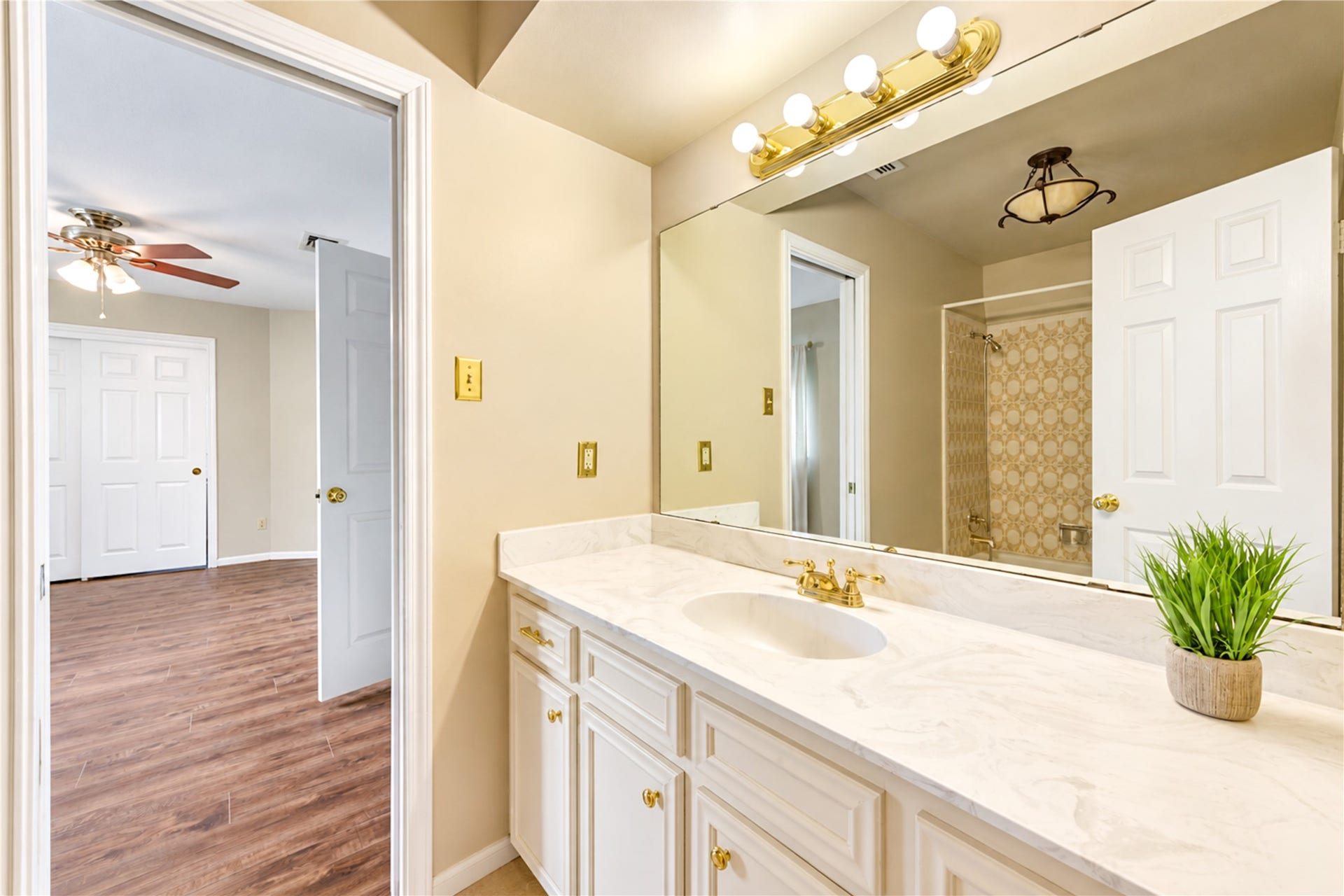 Elegant bathroom featuring a spacious marble countertop and vanity with gold fixtures, leading into a cozy bedroom with warm wood flooring and a ceiling fan.