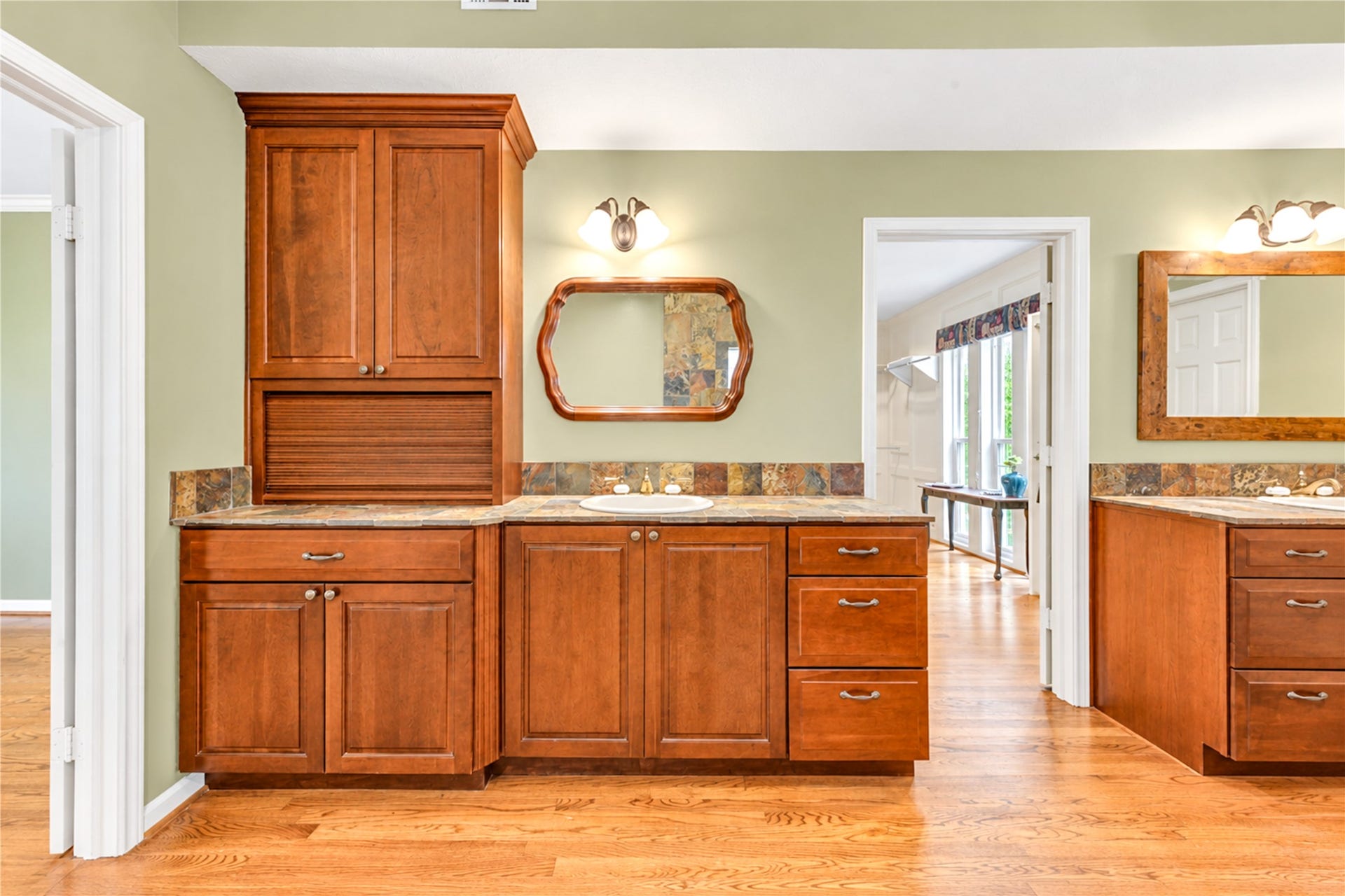 The large primary bathroom with rich wood cabinetry, stylish slate backsplash, and dual vanities, offering ample storage and classic charm.