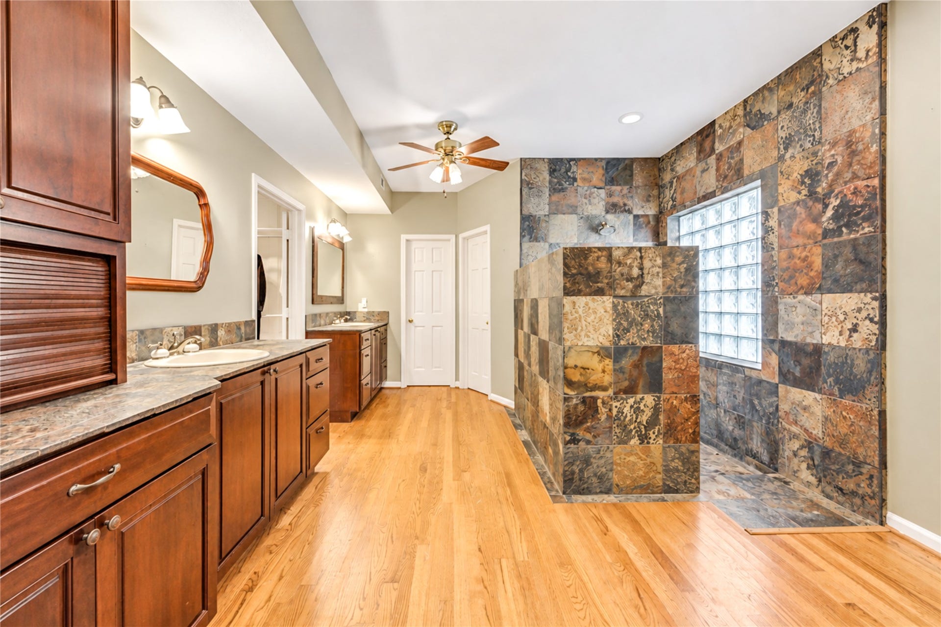 The primary bedroom features a spacious walk-in shower with slate tile, dual sinks with wooden cabinetry, and warm hardwood flooring, large water and linen closets.