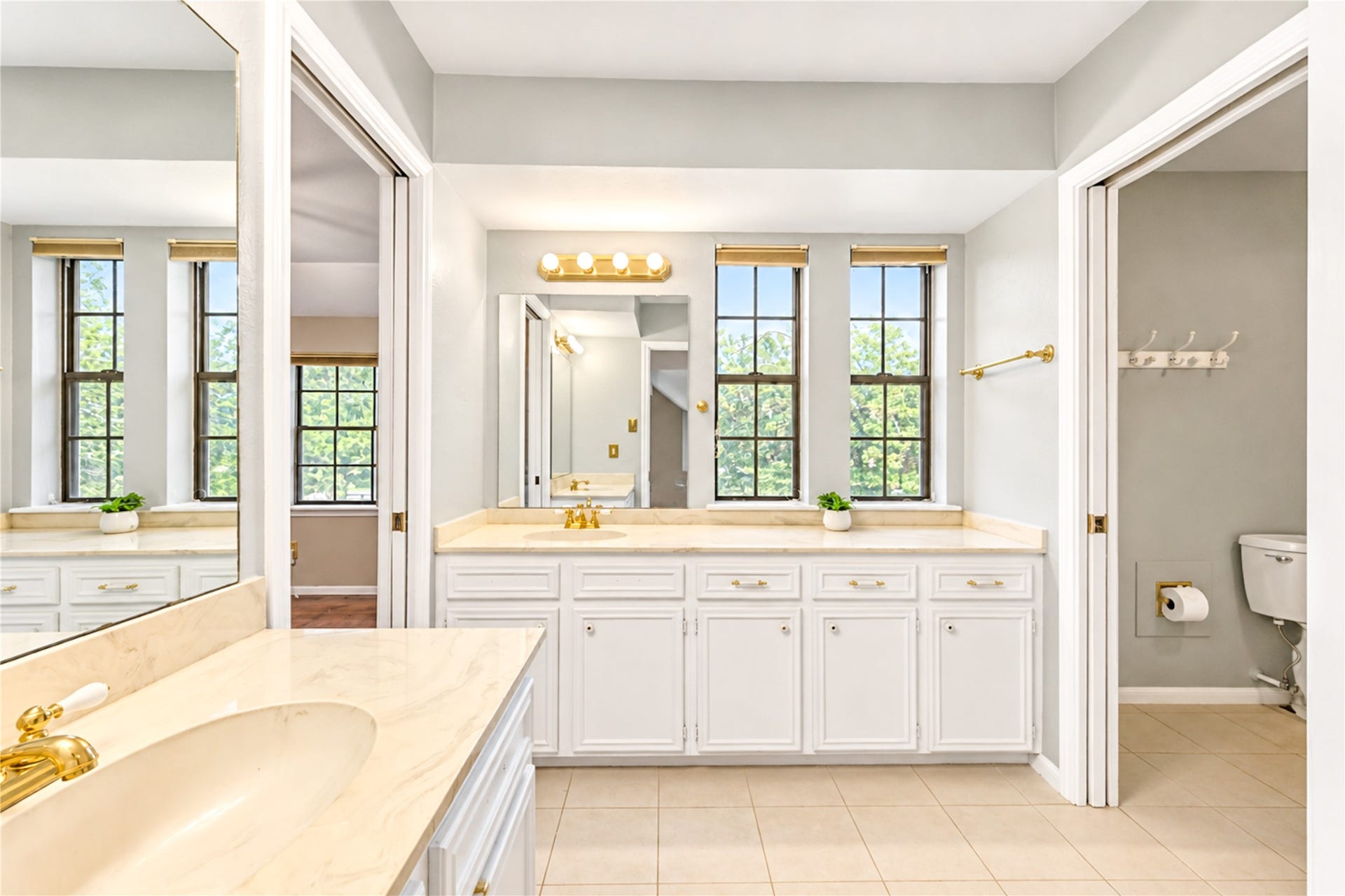 Bright and spacious bathroom featuring dual vanities with elegant gold fixtures, abundant natural light from large windows, and a separate toilet area for added privacy.