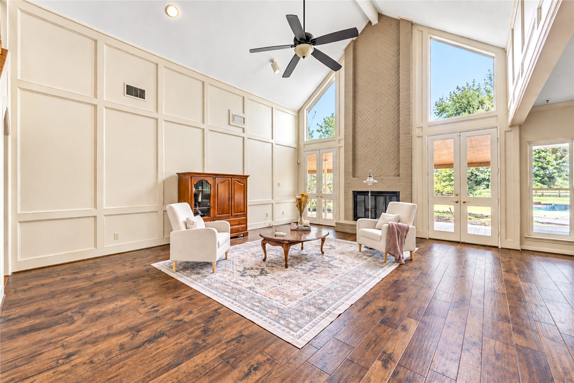 The spacious living room with soaring ceilings, elegant wood-look flooring, and abundant natural light from large windows, this living room features a cozy fireplace and glass doors leading to a serene outdoor pool and view.