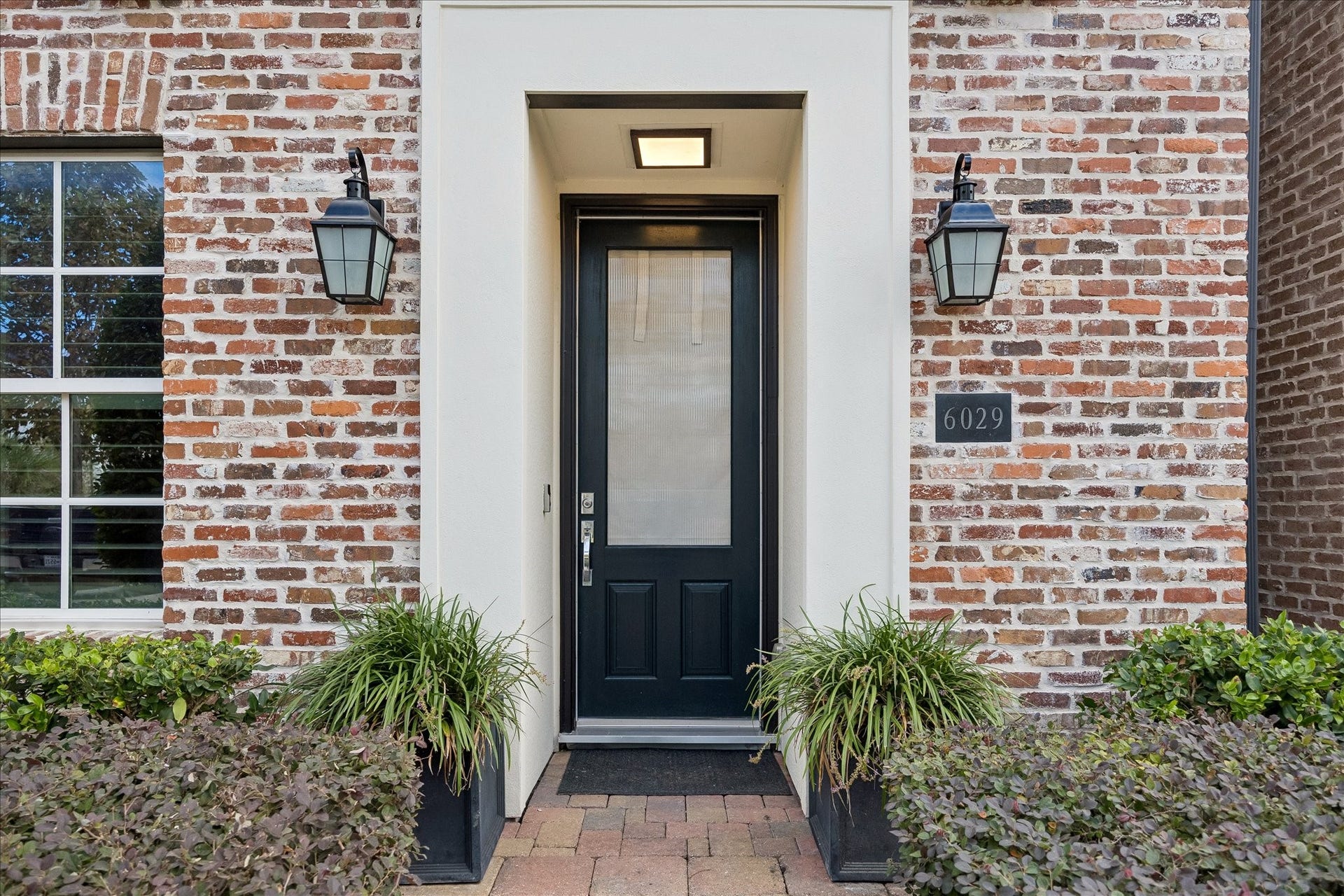6029 Matthew Oaks Place, Houston, TX 77007 Charming brick exterior with a classic black door and elegant lantern-style lighting, framed by lush greenery for a welcoming entrance.