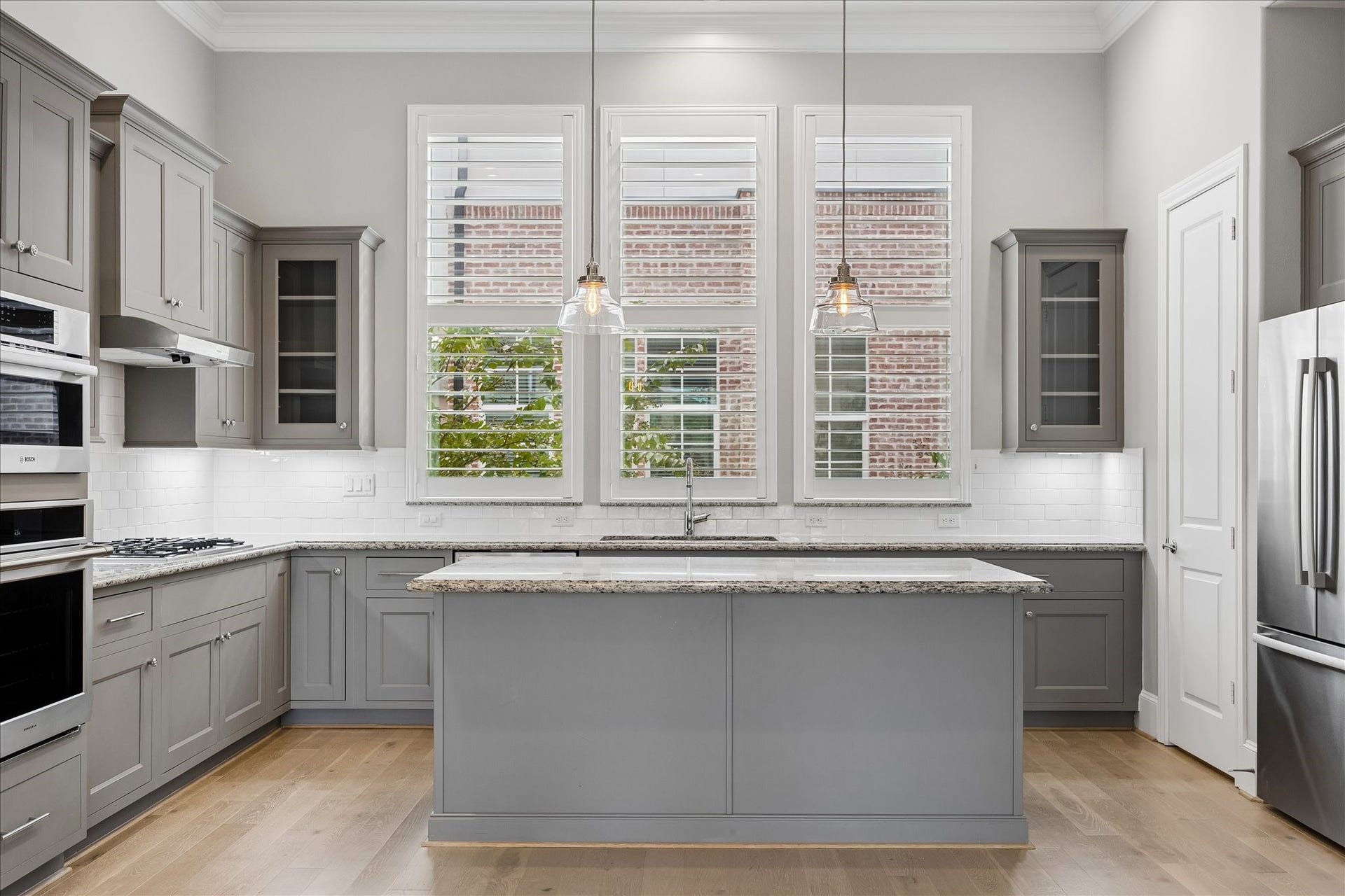 6029 Matthew Oaks Place, Houston, TX 77007 A modern kitchen featuring sleek gray cabinetry, stainless steel appliances, and a spacious island with pendant lighting, complemented by large windows with Plantation Shutters that offer ample natural light.