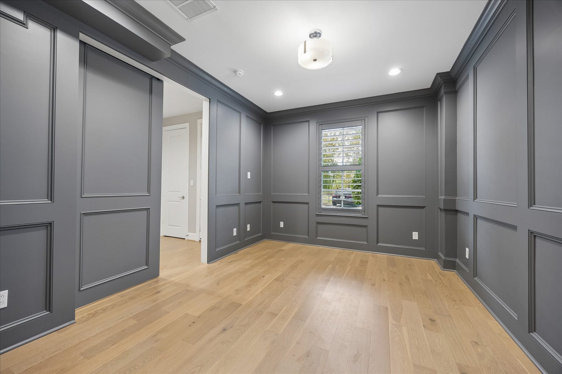 6029 Matthew Oaks Place, Houston, TX 77007 Elegant room with sophisticated dark gray wood paneled walls and warm hardwood floors, featuring recessed lighting and a modern ceiling fixture. A window with matching Plantation shutters offers natural light and a view. Perfect for an office or guest bedroom. It includes an en-suite full bath.