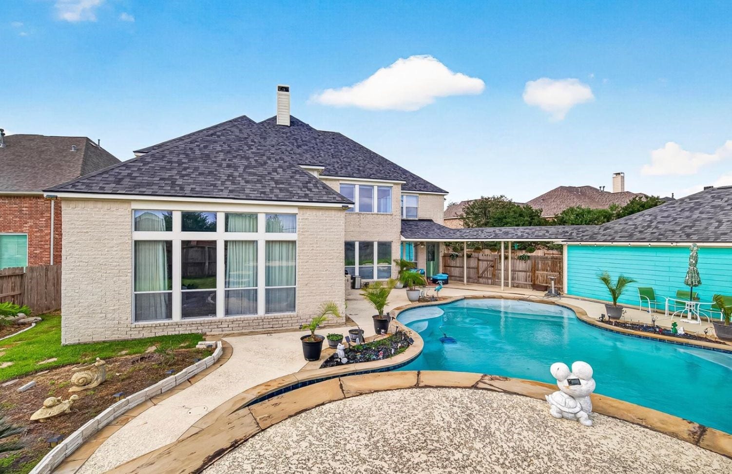 A fenced backyard with an in-ground pool and a covered patio offers a private outdoor retreat framed by green lawn and mulched planting beds. A large multi-pane window wall looks out over the pool area from the rear of the home.