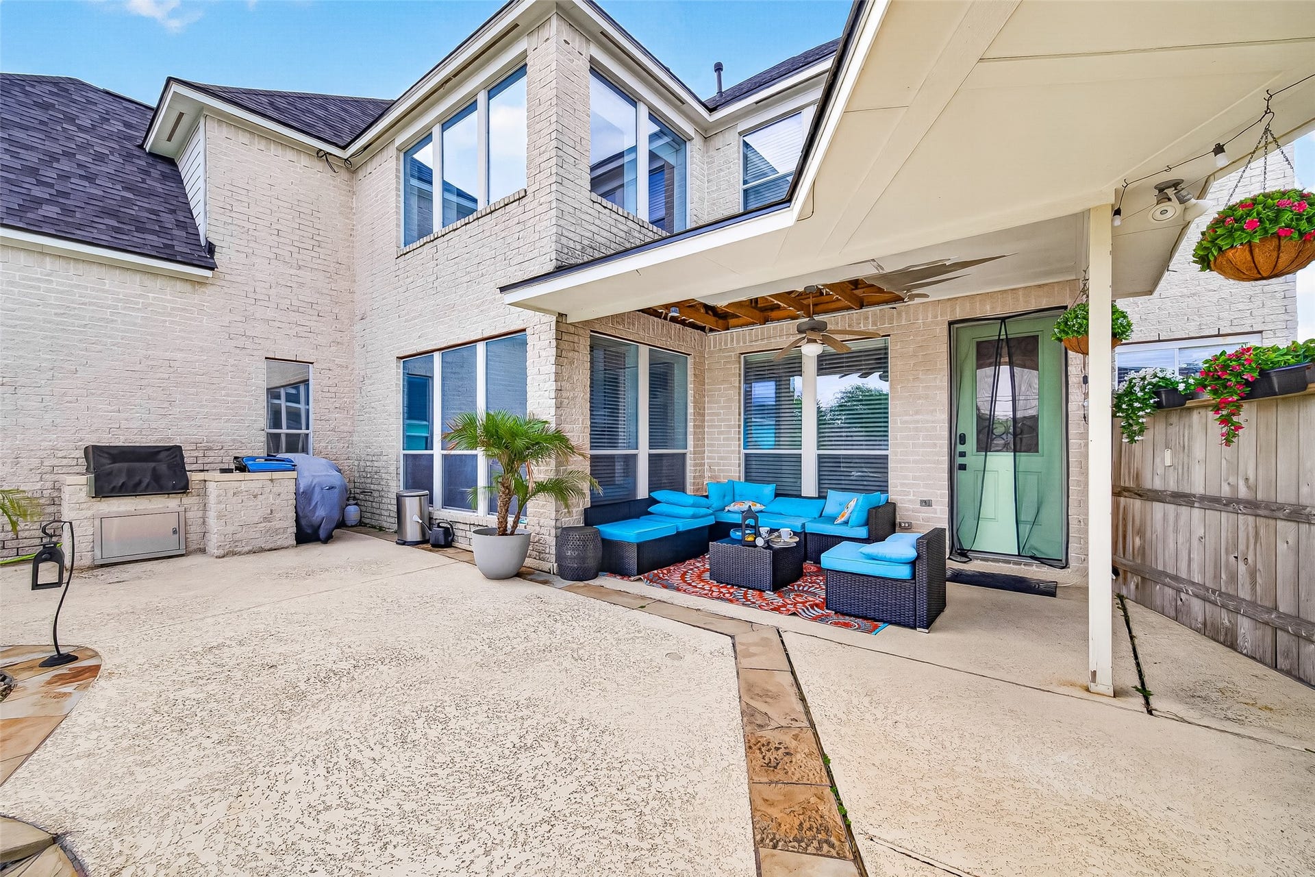 A covered patio extends the home's living space outdoors, framed by a wood pergola, a ceiling fan, and an outdoor kitchen with a stainless grill. An exposed-aggregate floor with a stone paver border and a fenced, landscaped yard complete the setting.