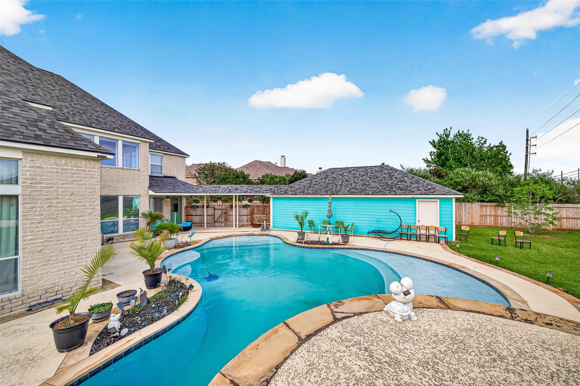 A freeform in-ground pool with flagstone coping anchors a fenced backyard framed by a covered patio and a detached outbuilding finished in teal siding. Mature trees and a broad lawn complete the private outdoor setting.
