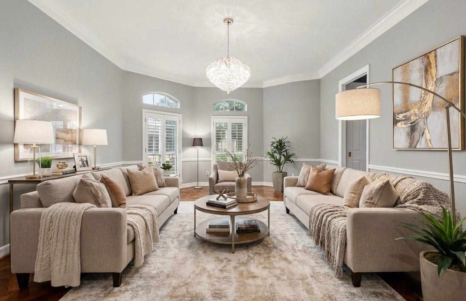 A formal living room defined by a tray ceiling with crown molding, white wainscoting, and paired French doors topped with arched transom windows, finished in hardwood flooring (Virtually staged).