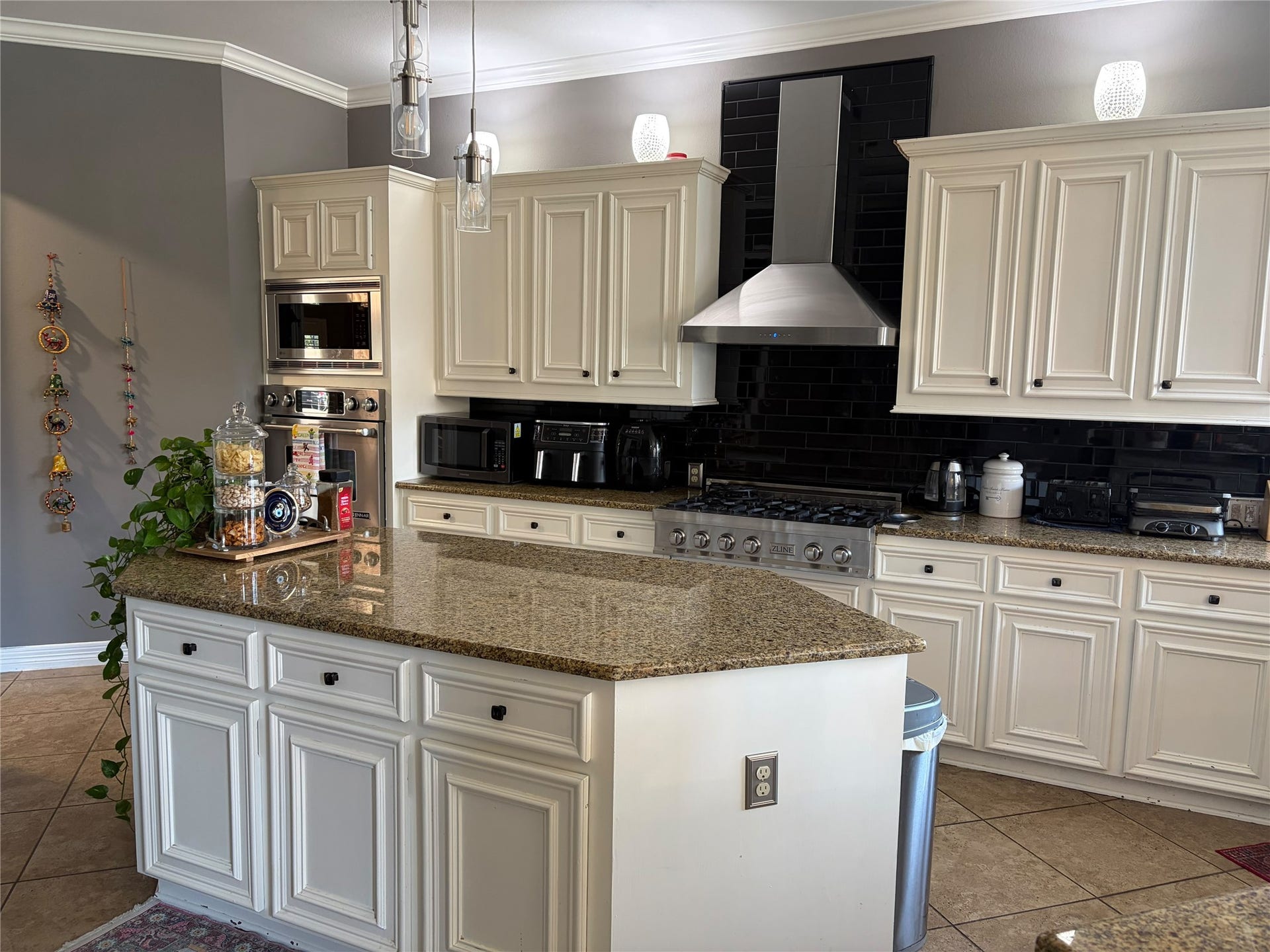 A spacious island kitchen anchors the main living area, featuring cream raised-panel cabinetry with crown molding, granite countertops, and a glossy black subway tile backsplash. Stainless steel appliances, including a gas range with a chimney-style hood, complete the space.(Original Picture)