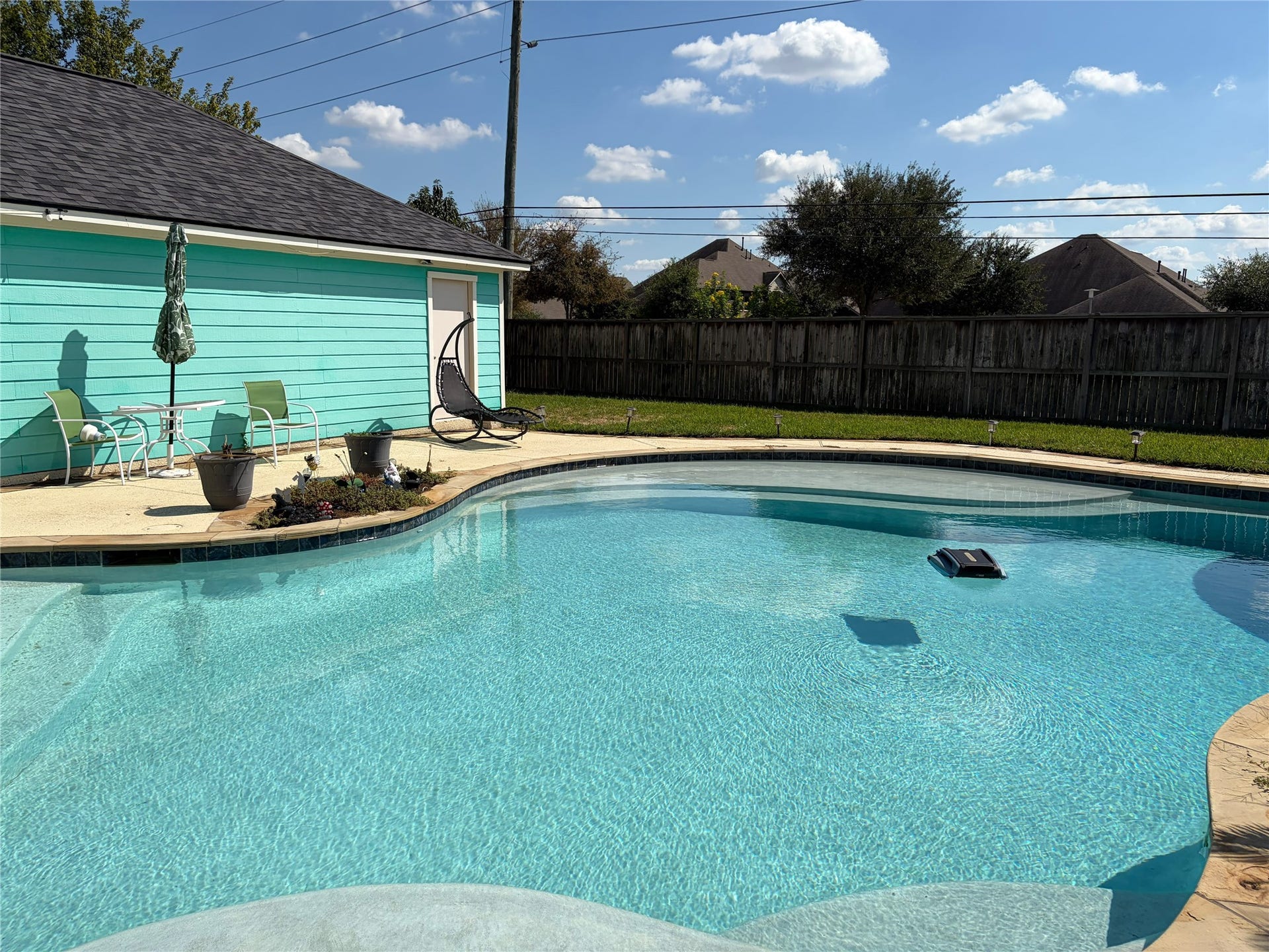 An in-ground pool with tan coping and a dark tile waterline anchors a fenced backyard, framed by a concrete patio and a strip of green lawn. A nearby seating area with a table, chairs, and a hanging chair completes the outdoor setting.