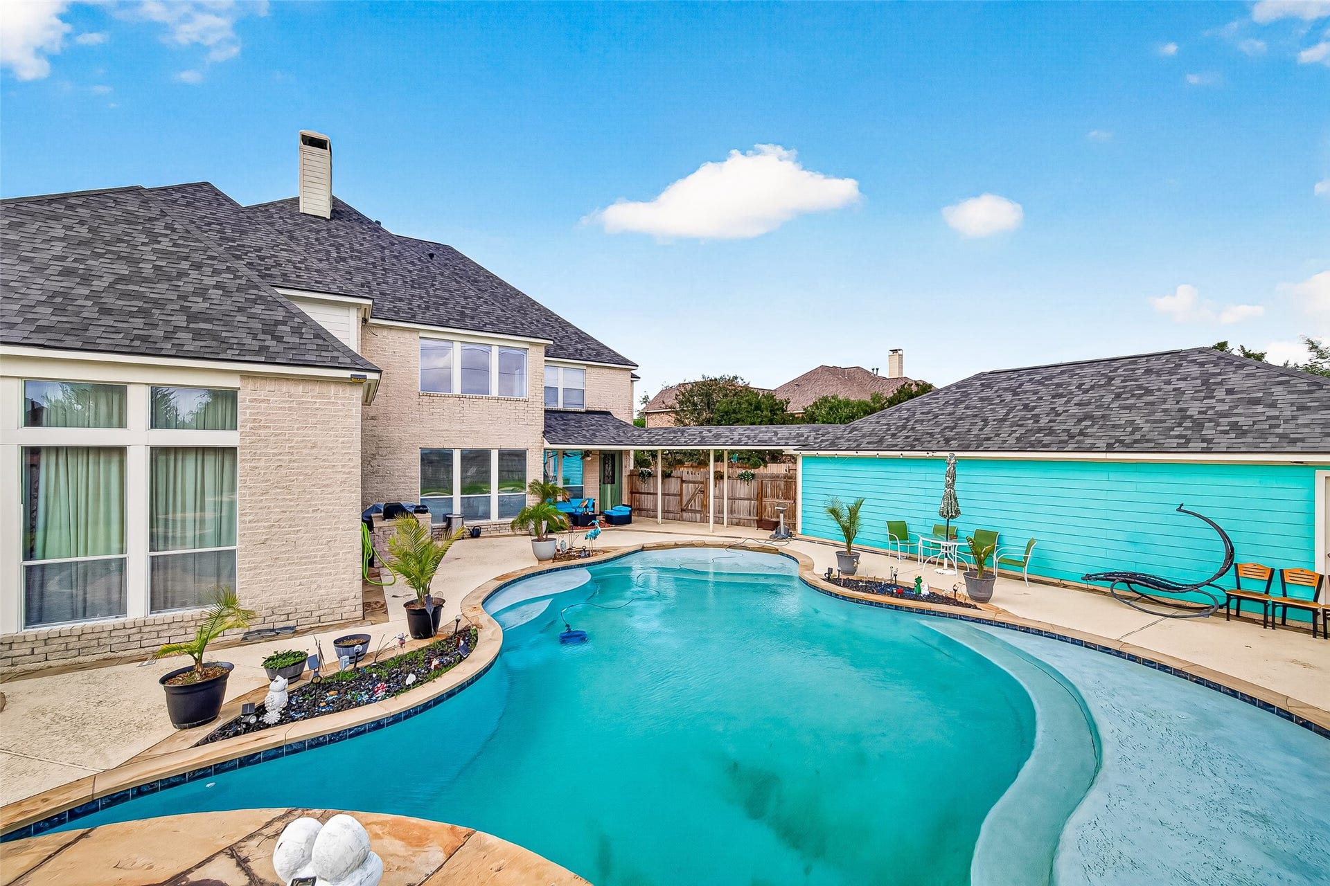 A freeform in-ground pool with a shallow tanning ledge anchors the rear patio, framed by a covered pergola and a turquoise-sided outbuilding. A two-story brick elevation with tall multi-pane windows overlooks the fenced and landscaped yard.