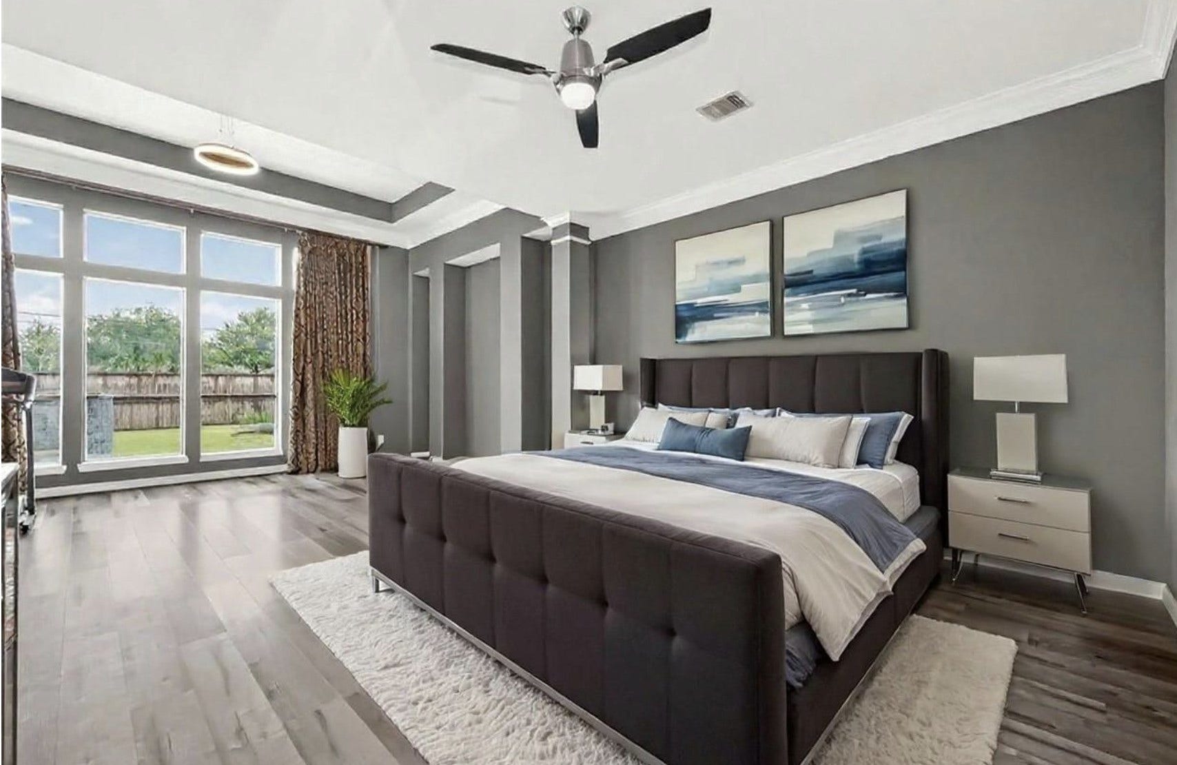 A primary bedroom with a tray ceiling and crown molding, framed by a wall of windows with patterned drapery that fills the space with natural light. (Virtually Staged).