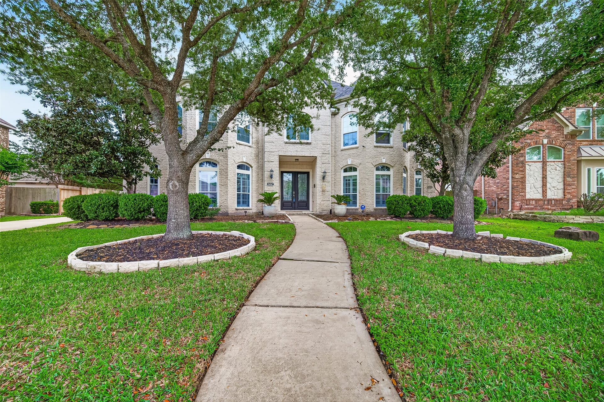 A cut-stone veneer facade with multiple gables and a recessed covered entry establishes a refined first impression at the front elevation. Mature shade trees, a manicured lawn, and a curved concrete walkway lead to glass-paneled double doors flanked by wall sconces (Original Picture).