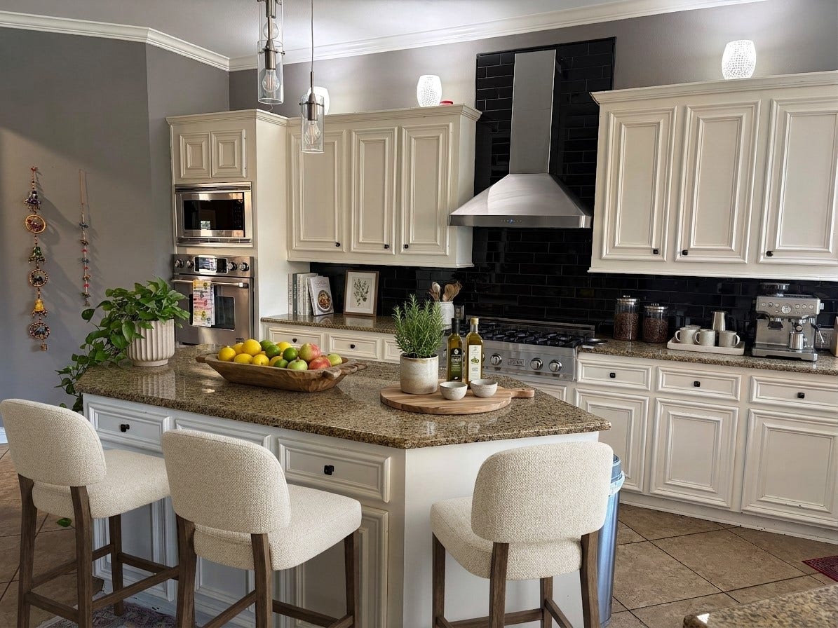 An island kitchen pairs cream raised-panel cabinetry and granite countertops with a black subway-tile backsplash and a full stainless steel appliance suite. Glass pendant lights hang above the island, where upholstered bar seating and a wood-framed window define the space.(Virtually Staged)