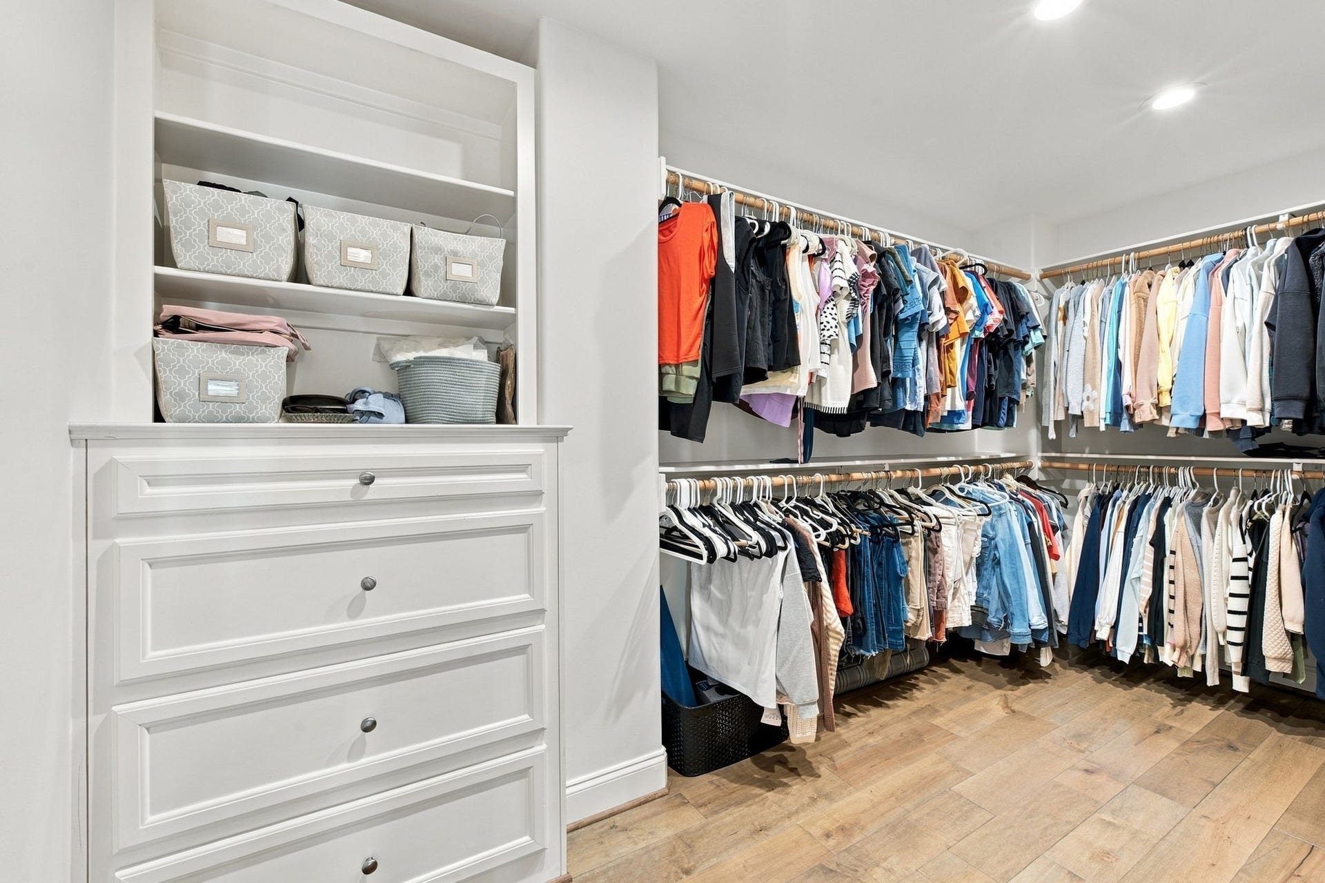 Spacious primary walk-in closet featuring custom built-in drawers, open shelving, double hanging rods & hardwood floors for exceptional organization & storage.