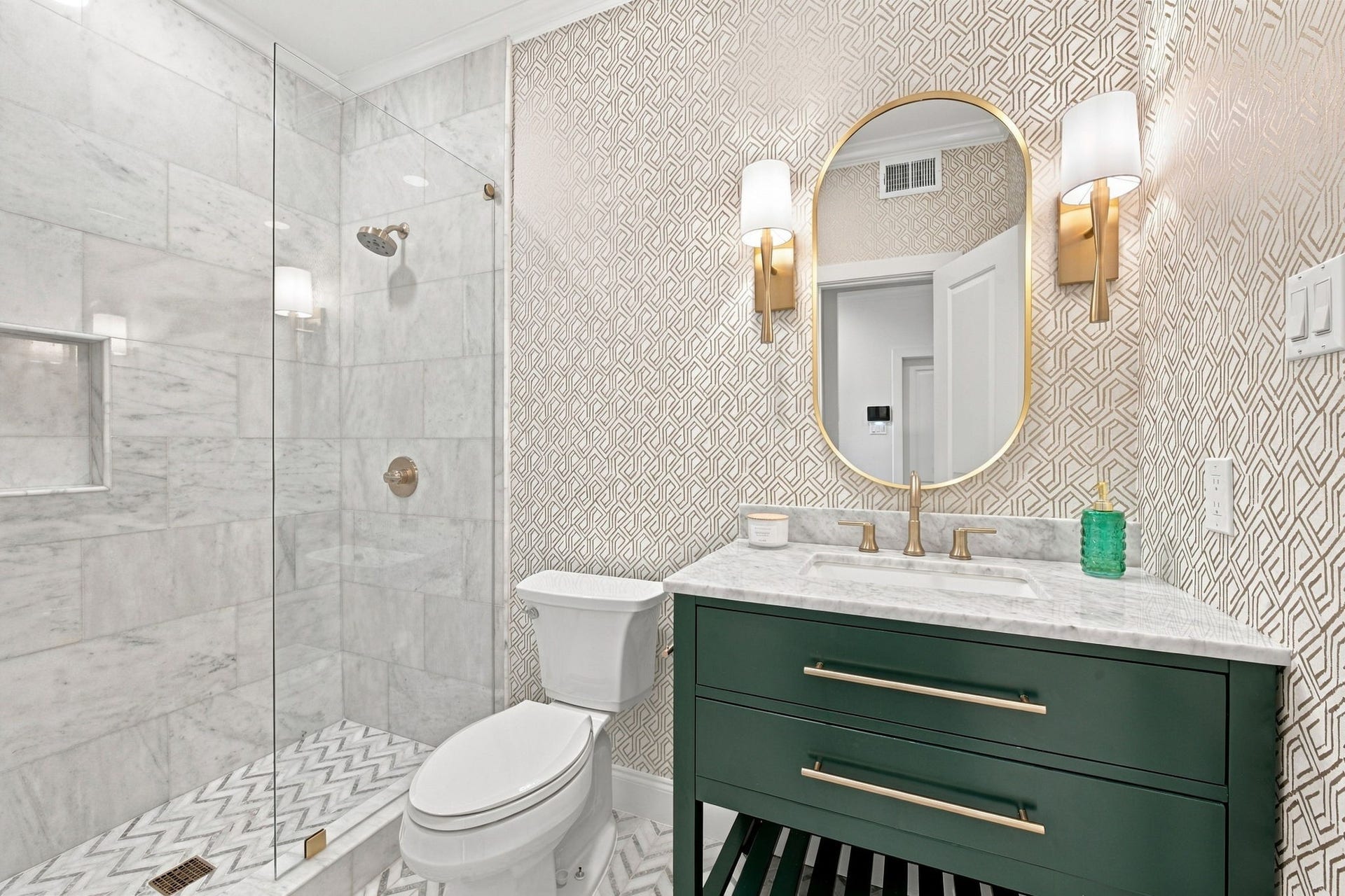 Beautifully updated full bath featuring a glass-enclosed walk-in shower w/marble tile & built-in niche, striking geometric wallpaper, marble countertop, rich green vanity & elegant gold fixtures & accents.