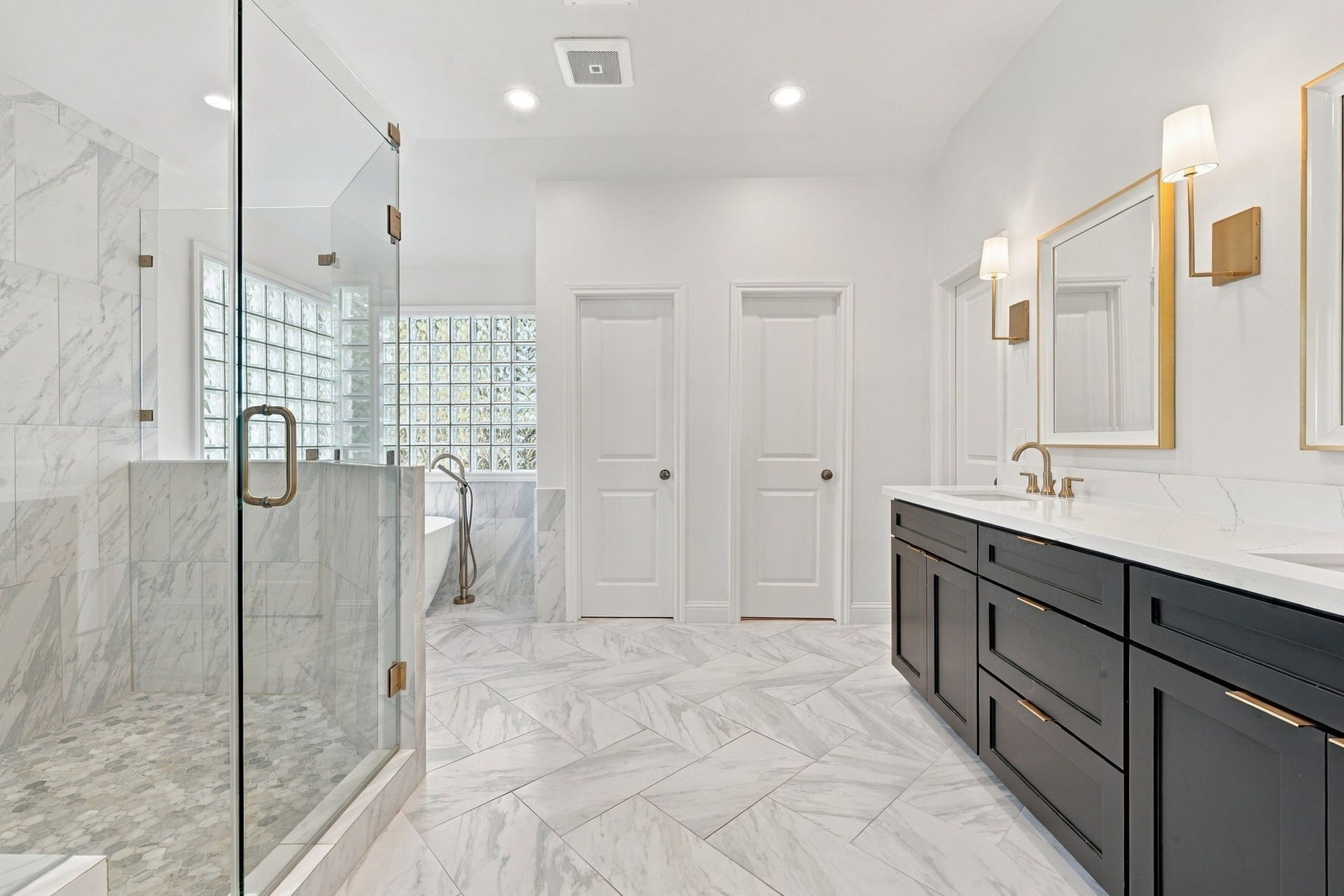 Beautifully appointed primary bath featuring marble-look tile floors, a spacious glass-enclosed shower, freestanding soaking tub & dual-sink vanity w/dark cabinetry, gold accents & elegant lighting.