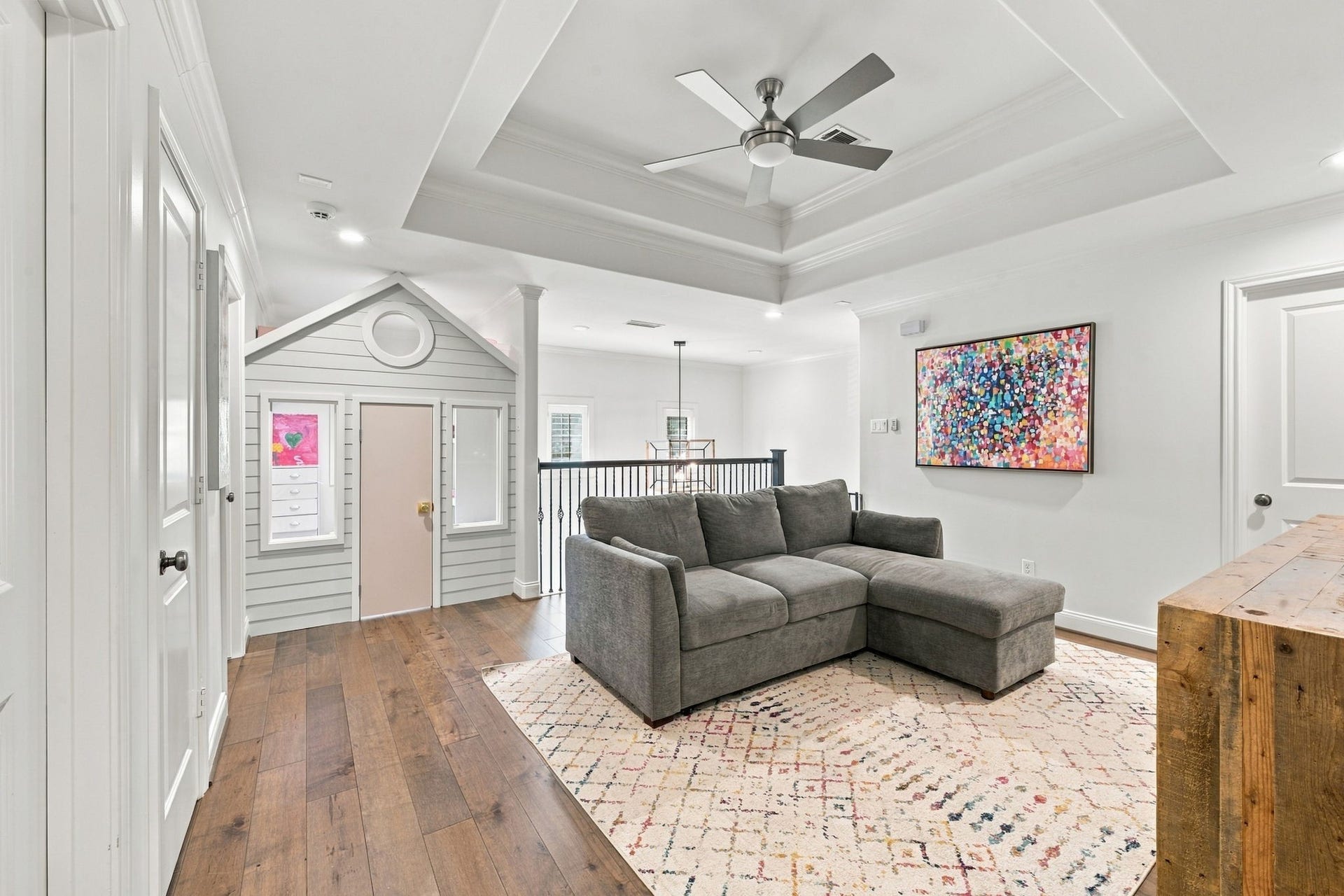 Wonderful view of the upstairs game room showcasing the custom built-in dollhouse, beautiful tray ceiling, hardwood floors & open railing overlooking the entry. A fantastic space for family gatherings, entertainment & imaginative play!