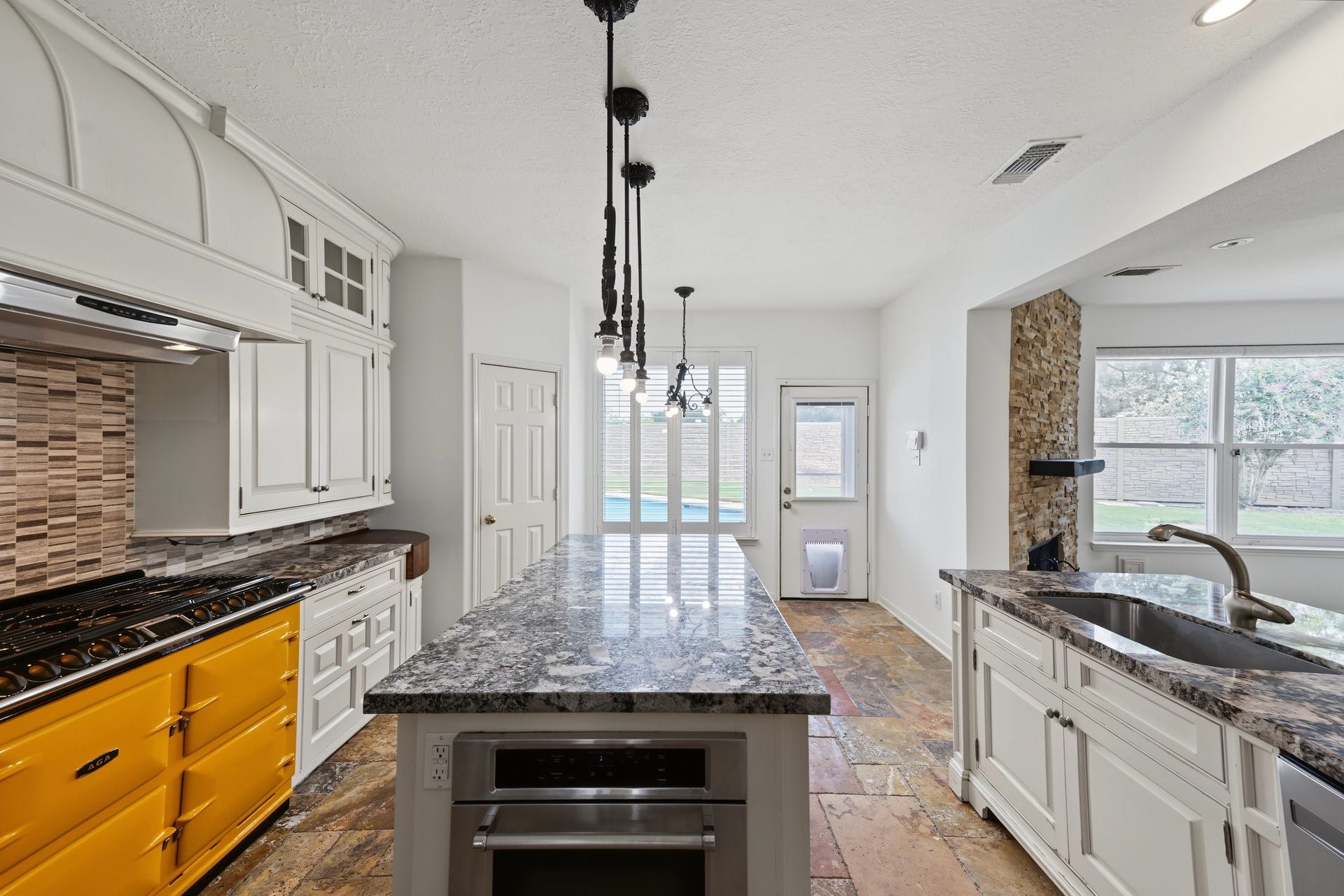 Stunning kitchen with a bold yellow AGA range, elegant granite countertops, and a spacious island, complemented by sleek pendant lighting and a vibrant backsplash. Add your own pendant shades to reflect your style.