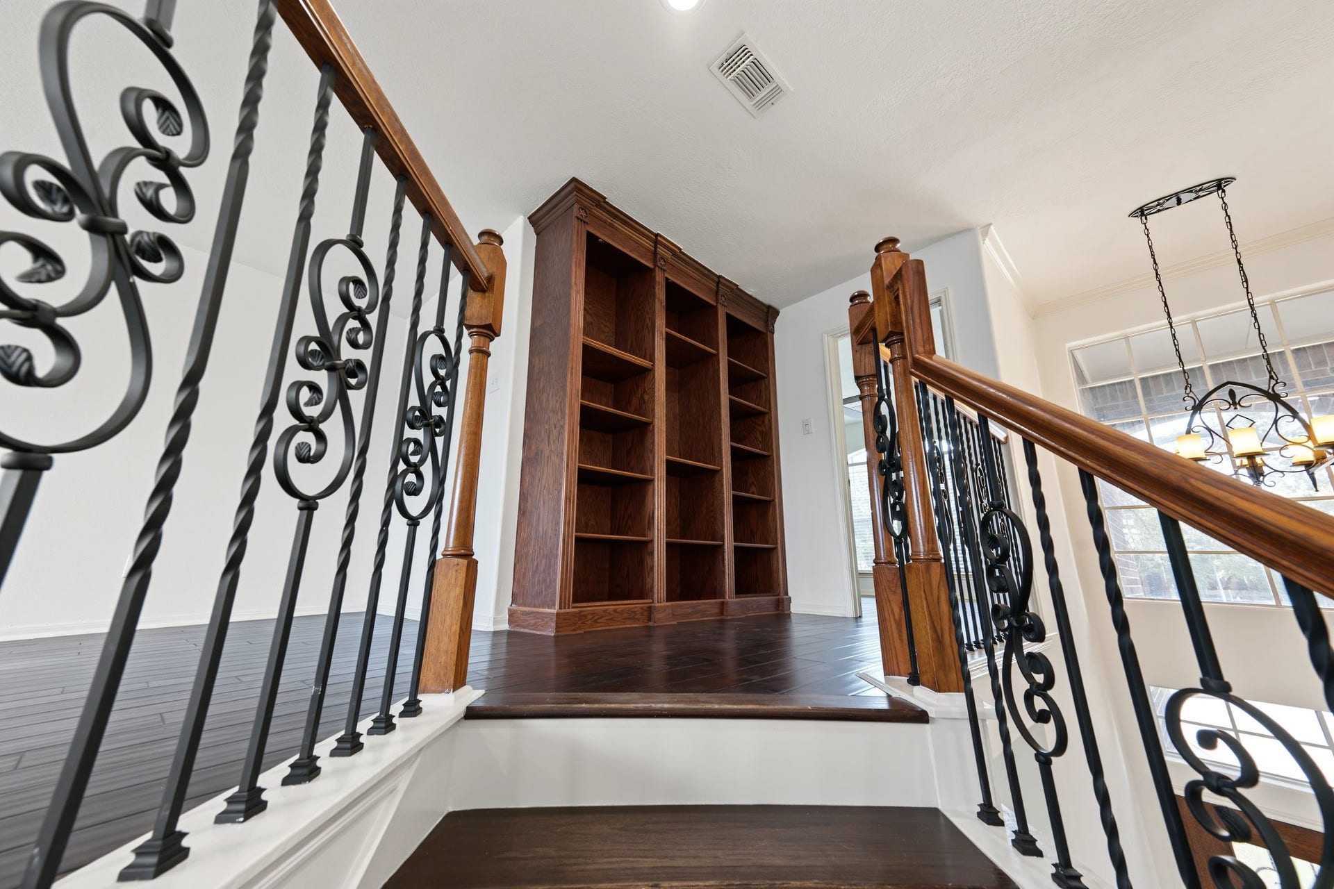 Staircase with ornate wrought iron railings leads to a cozy landing area featuring a built-in wooden bookshelf, enhanced by natural light from large windows. A great place for your library!