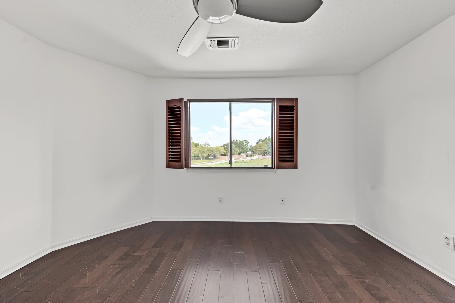 Bright and spacious room featuring elegant dark hardwood floors, fresh white walls, and a modern ceiling fan, with a large window offering scenic views of the backyard. Bright and cheery.