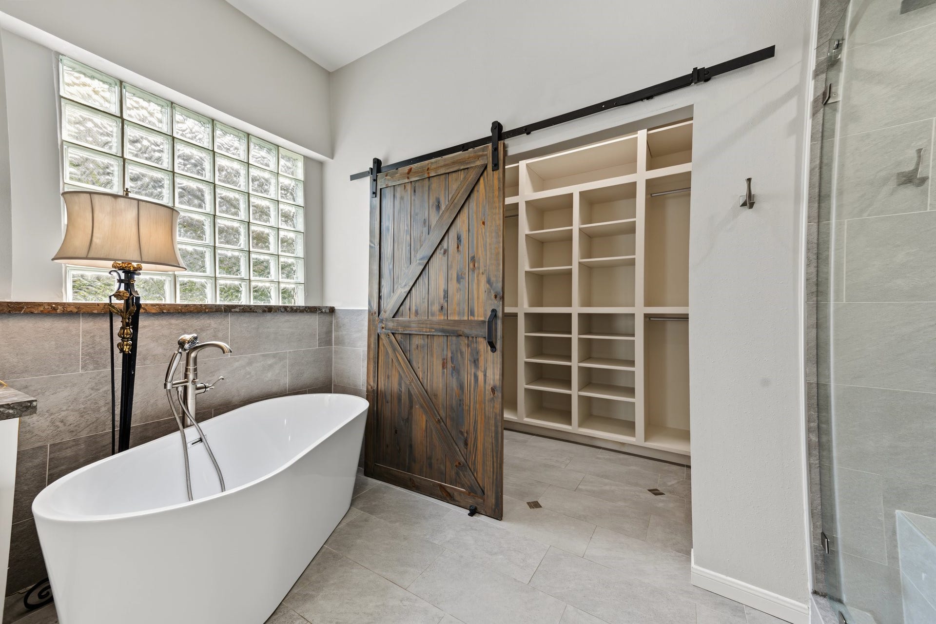 Luxurious primary bathroom featuring a freestanding soaking tub, elegant glass block window, and a rustic sliding barn door leading to a spacious walk-in closet and custom built ins. Look how many shoes you can store!