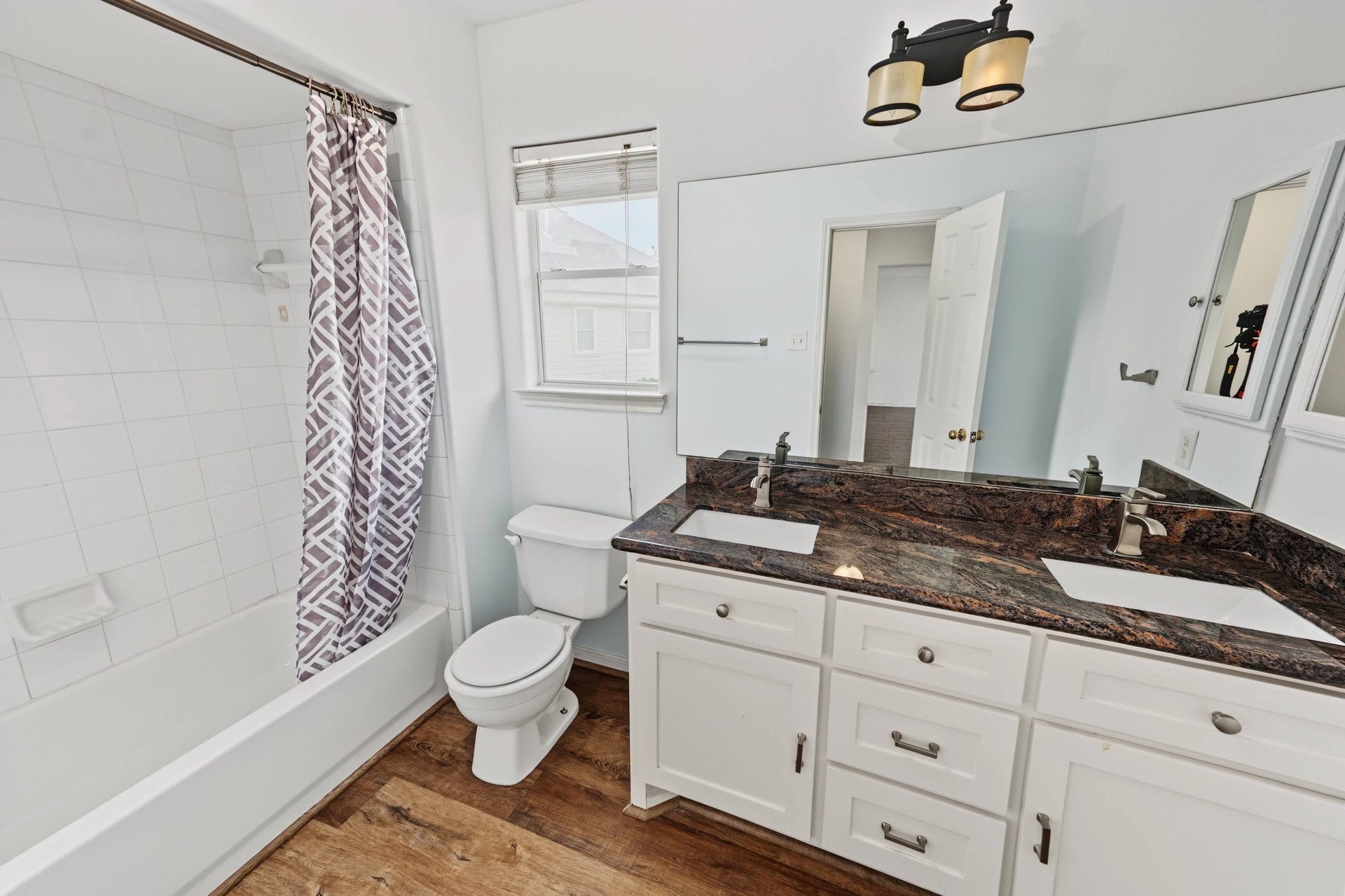Bright and inviting bathroom featuring a double vanity with granite countertops, a large mirror, and a shower-tub combo.