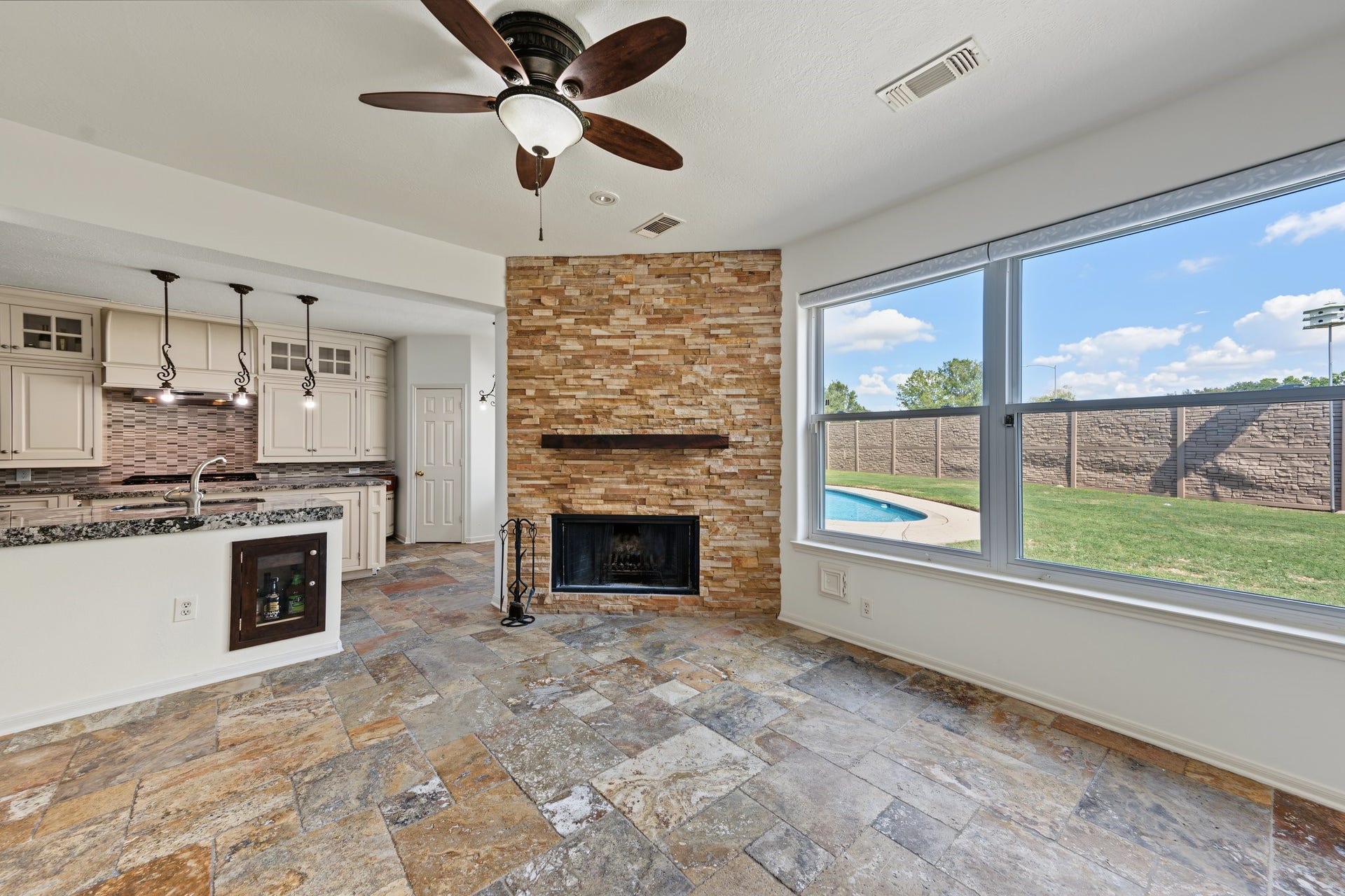 The living room is illuminated with natural light through large windows. A beautiful stone fireplace compliments the view of the backyard pool. The open space concept connects the living room to a modern kitchen, equipped with granite countertops and stylish pendant lighting.
