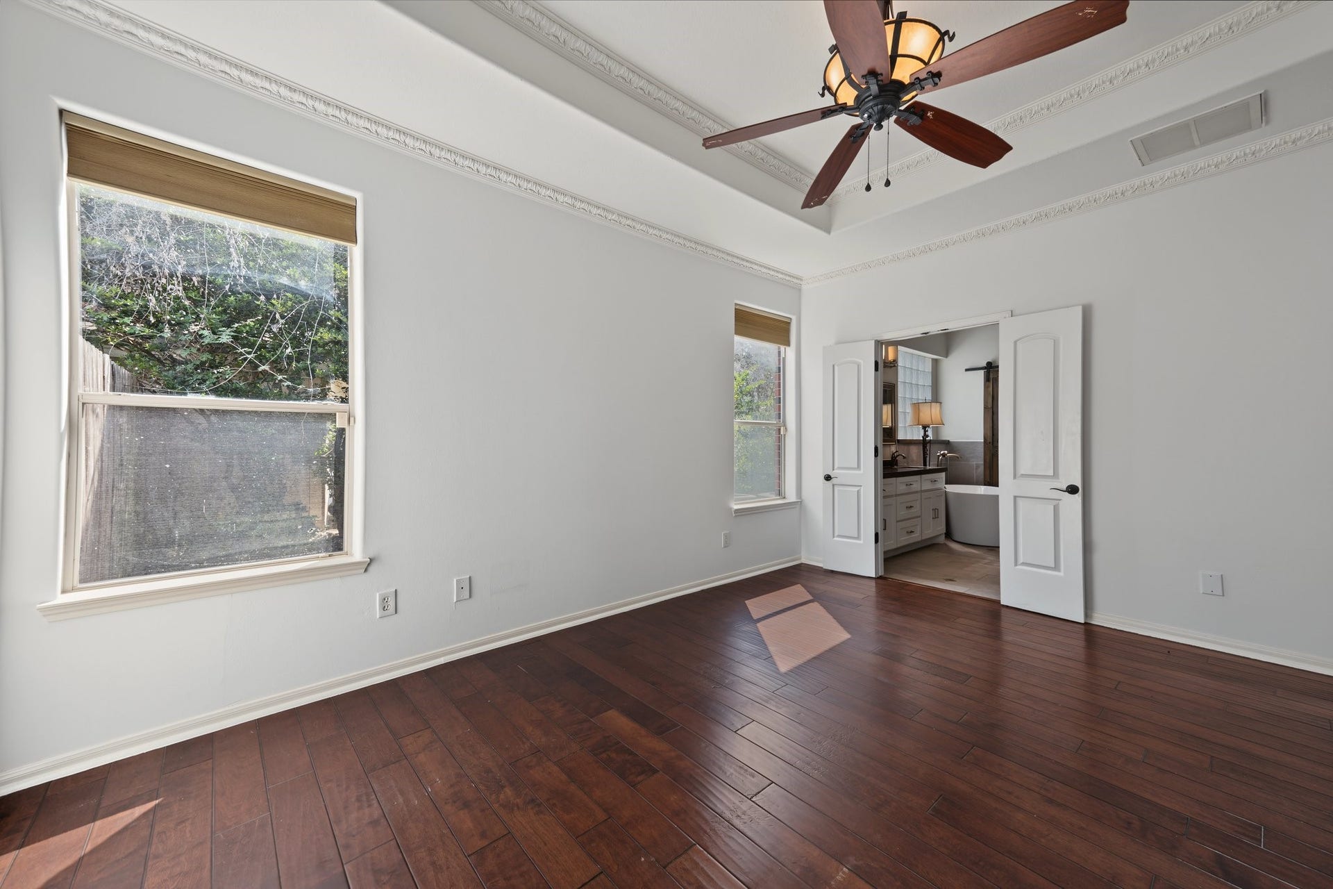 The primary bedroom featuring rich hardwood floors, elegant tray ceiling with a stylish ceiling fan, with direct access to a modern ensuite bathroom through double doors. Ready to move in!