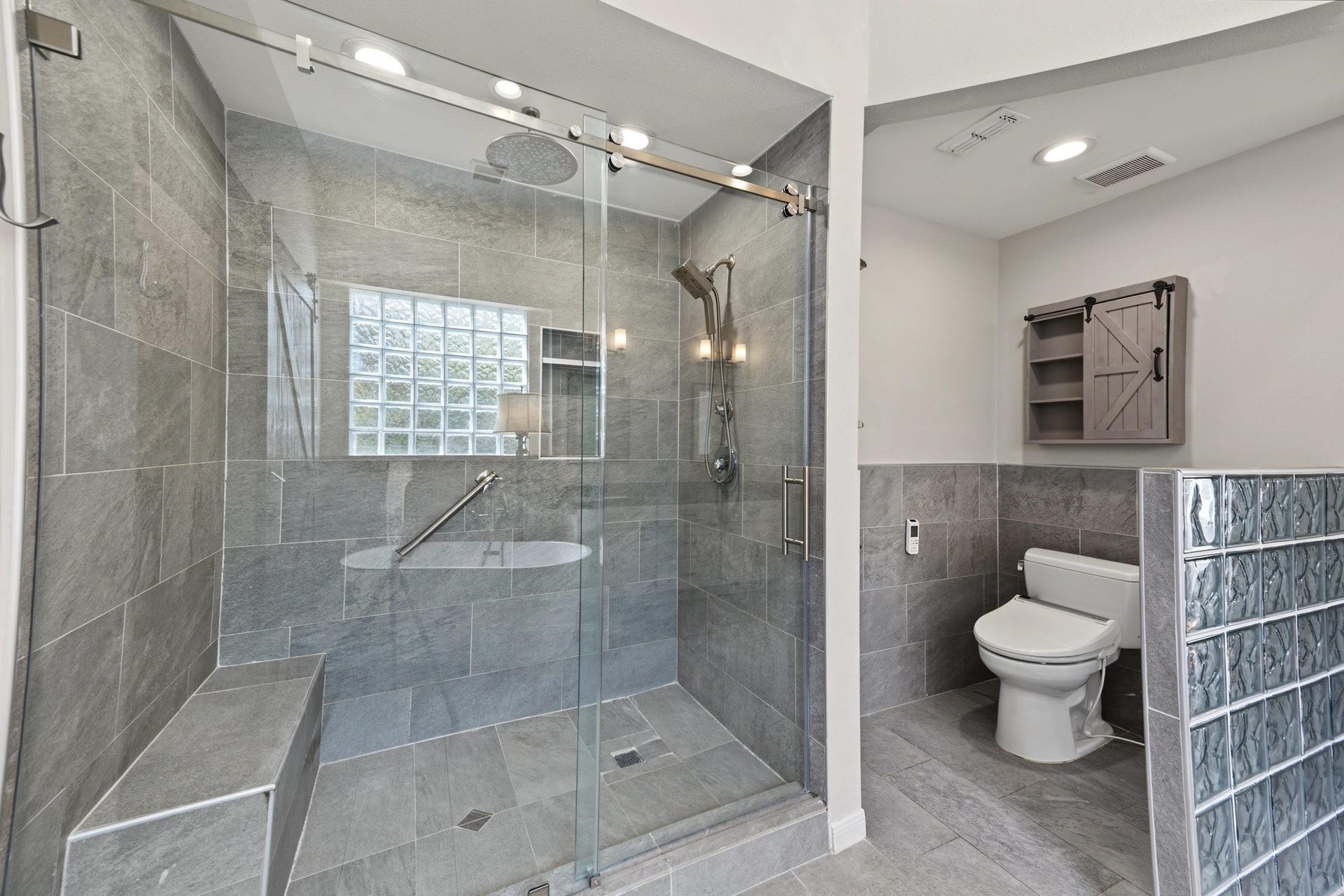 Modern bathroom featuring a spacious glass-enclosed shower with sleek gray tiles and a built-in bench, complemented by a stylish toilet area with frosted glass block accents. Elegant!
