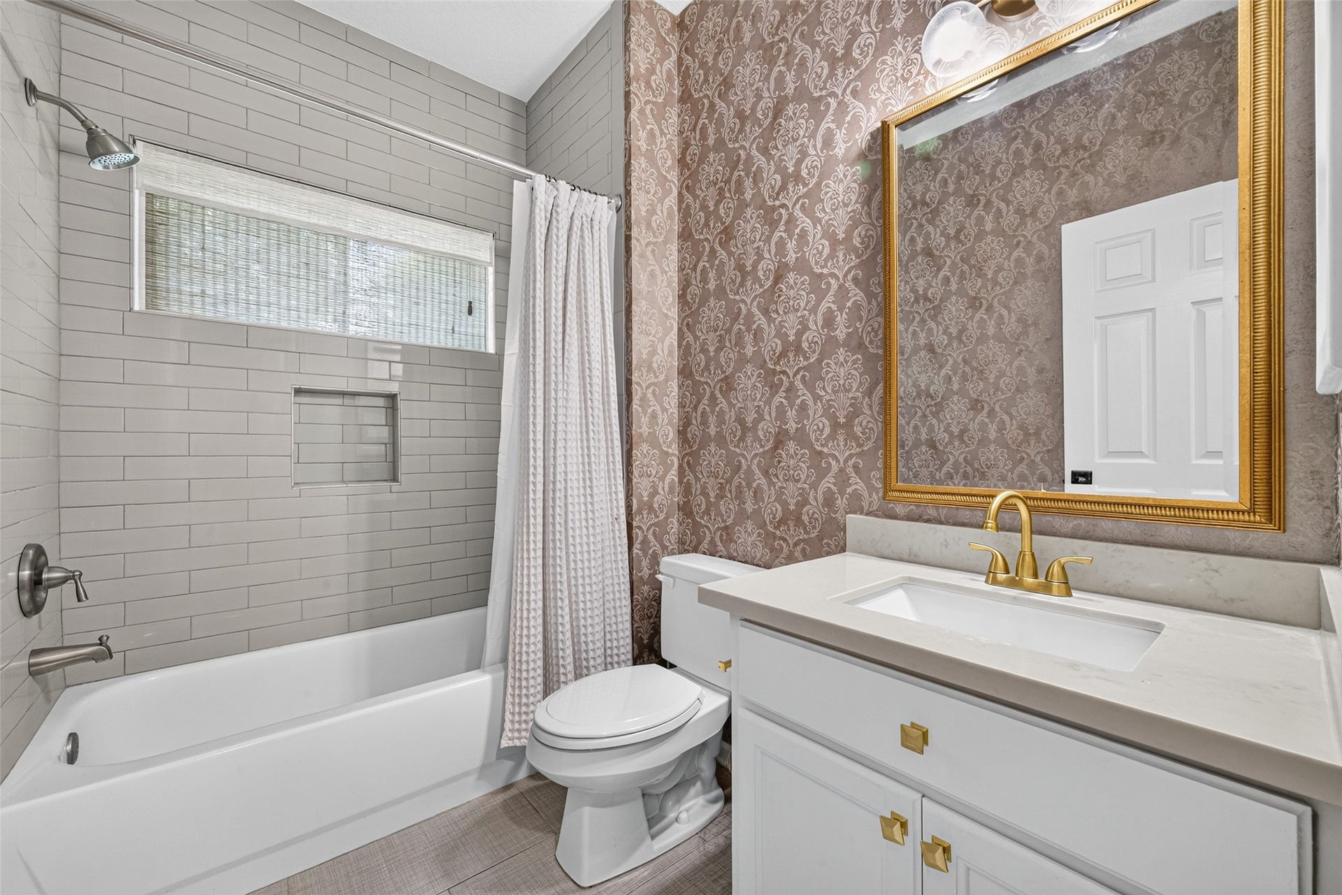 7707 Stone Arbor Drive, Sugar Land, TX 77479 A BEAUTIFULLY APPOINTED 1ST FLOOR BATH! Conveniently located near the Study/Bedroom #4, this full bath features a tub/shower combination surrounded by white subway tile extending to the ceiling, a recessed shower niche & a window welcoming natural light. The white vanity is complemented by a light-colored countertop, brushed gold fixtures, coordinating cabinet hardware & a decorative framed mirror. Patterned wallpaper adds a touch of personality, creating a stylish space for everyday use or guests.
