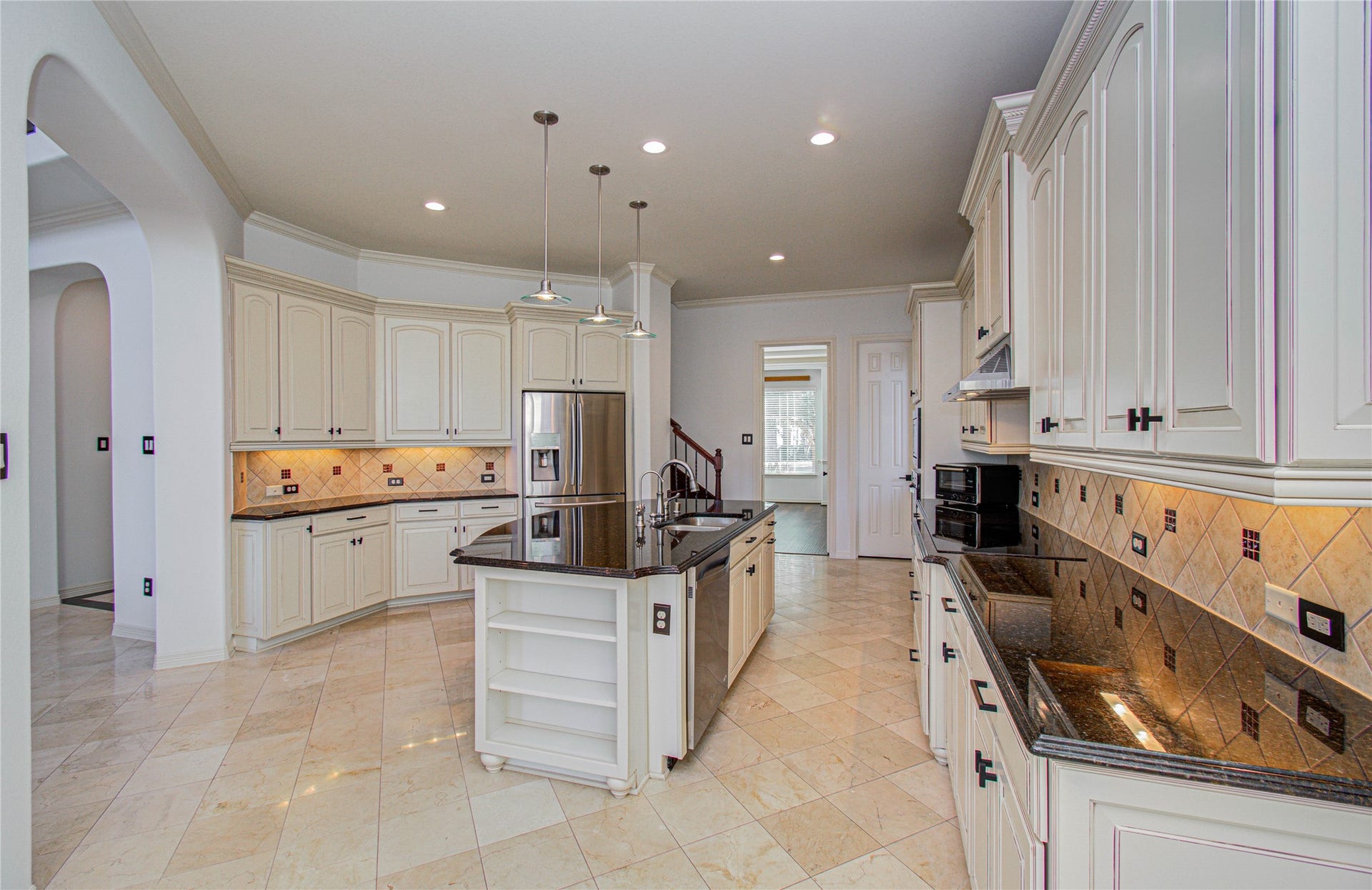 spacious, elegant kitchen with granite countertops, an island with pendant lighting, and stainless steel appliances. The room is open and bright, with light tile flooring and cream-colored cabinetry. Adjacent is a wooden staircase leading to the upper floor, adding warmth and sophistication to the space.