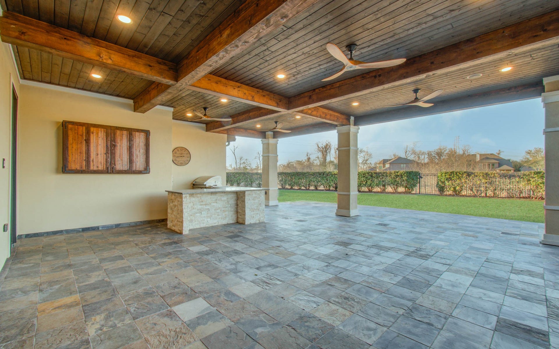 This photo showcases a spacious, covered patio with a stylish tiled floor, wood-paneled ceiling, ceiling fans, and built-in outdoor kitchen, overlooking a neatly manicured lawn and hedges. Ideal for entertaining and relaxation.