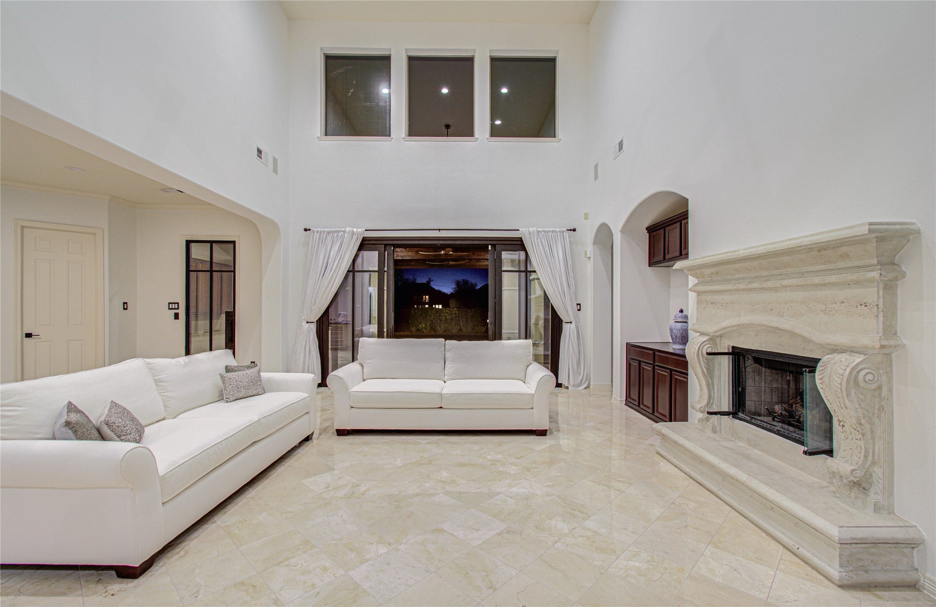 This spacious living room features high ceilings, elegant marble floors, and a grand fireplace. Large windows provide natural light, and the open layout includes comfortable seating and classic decor, creating a luxurious and inviting atmosphere.