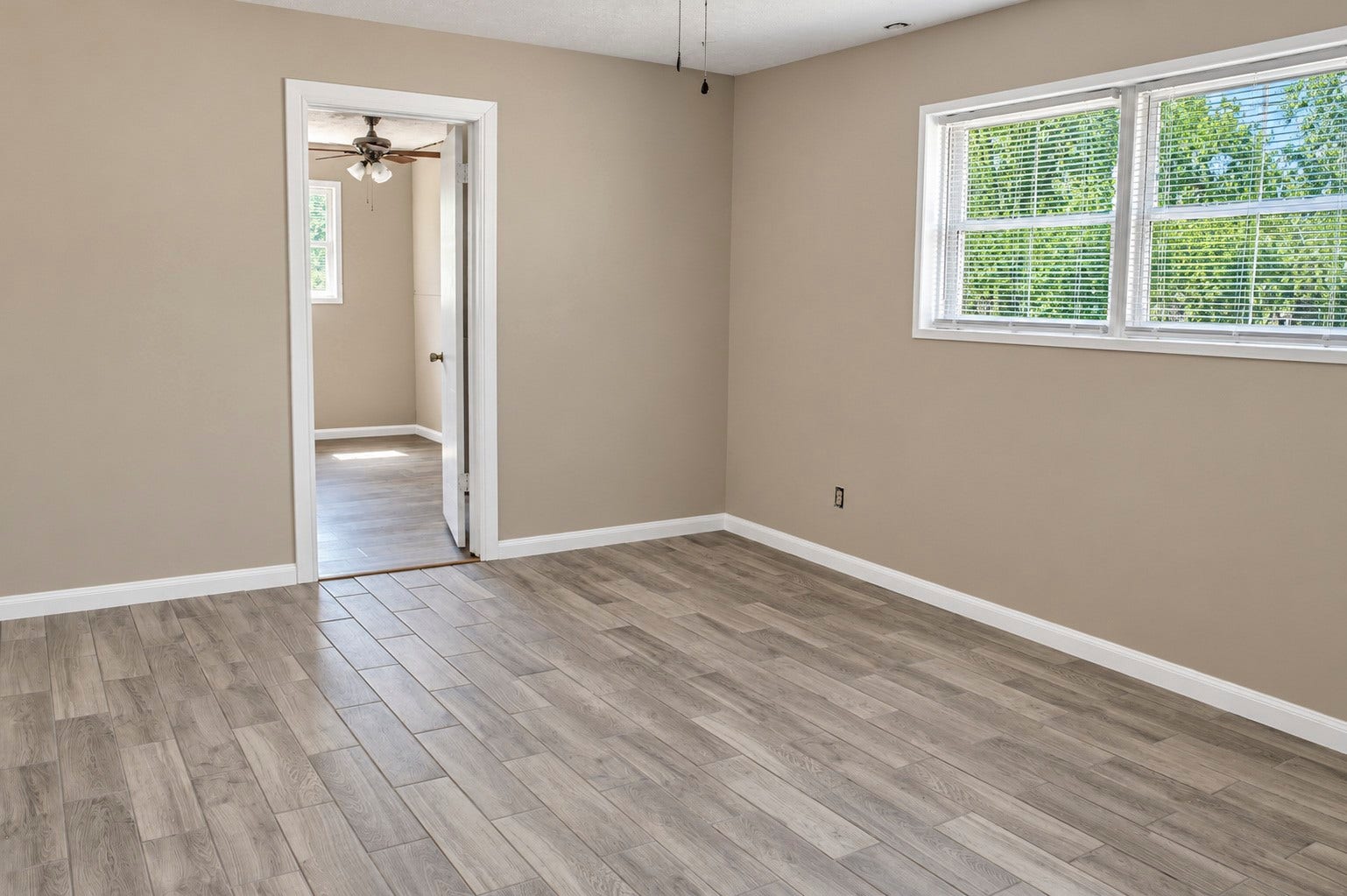 Spacious bedroom with wood-look tile flooring and abundant natural light leading into laundry room with a ceiling fan.