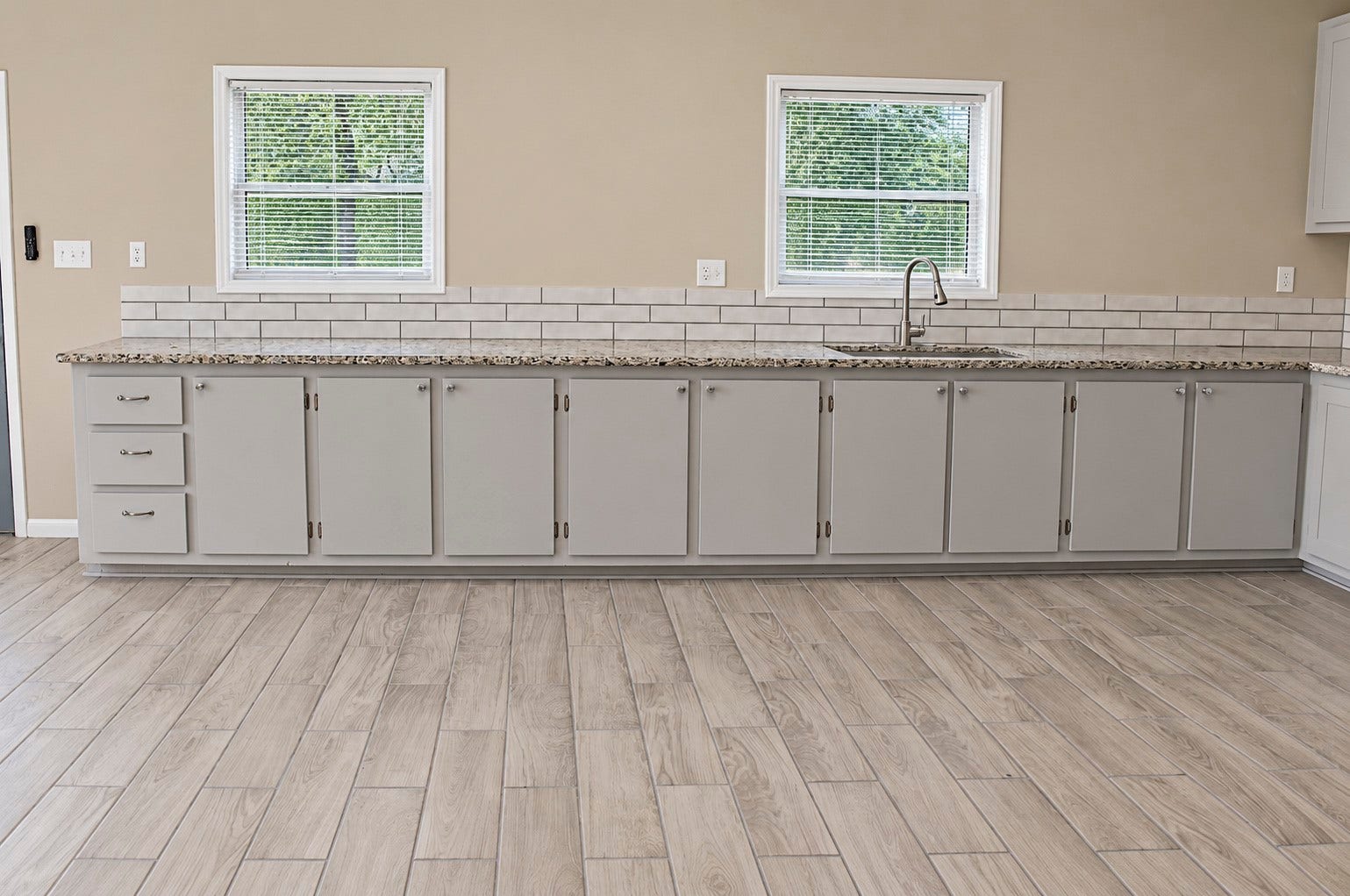 Spacious kitchen with elegant granite countertops, sleek light gray cabinetry, and a modern subway tile backsplash, complemented by warm wood-look tile flooring.
