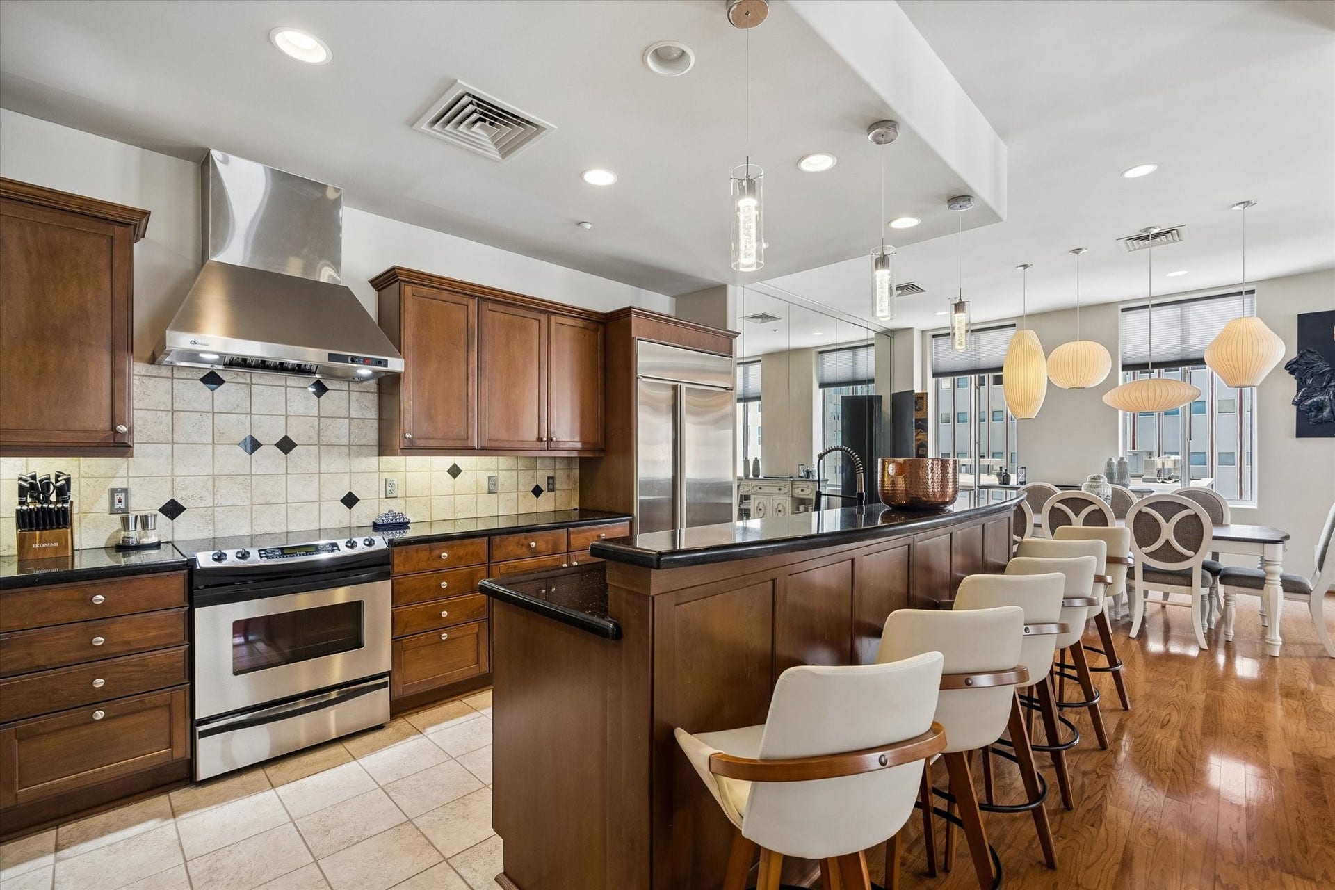 914 Main Street, #801, Houston, TX 77002 Elegant kitchen with rich wood cabinetry, stainless steel appliances, and a stylish tile backsplash. The open layout includes a breakfast bar with chic seating, flowing seamlessly into a bright dining area enhanced by modern pendant lighting.